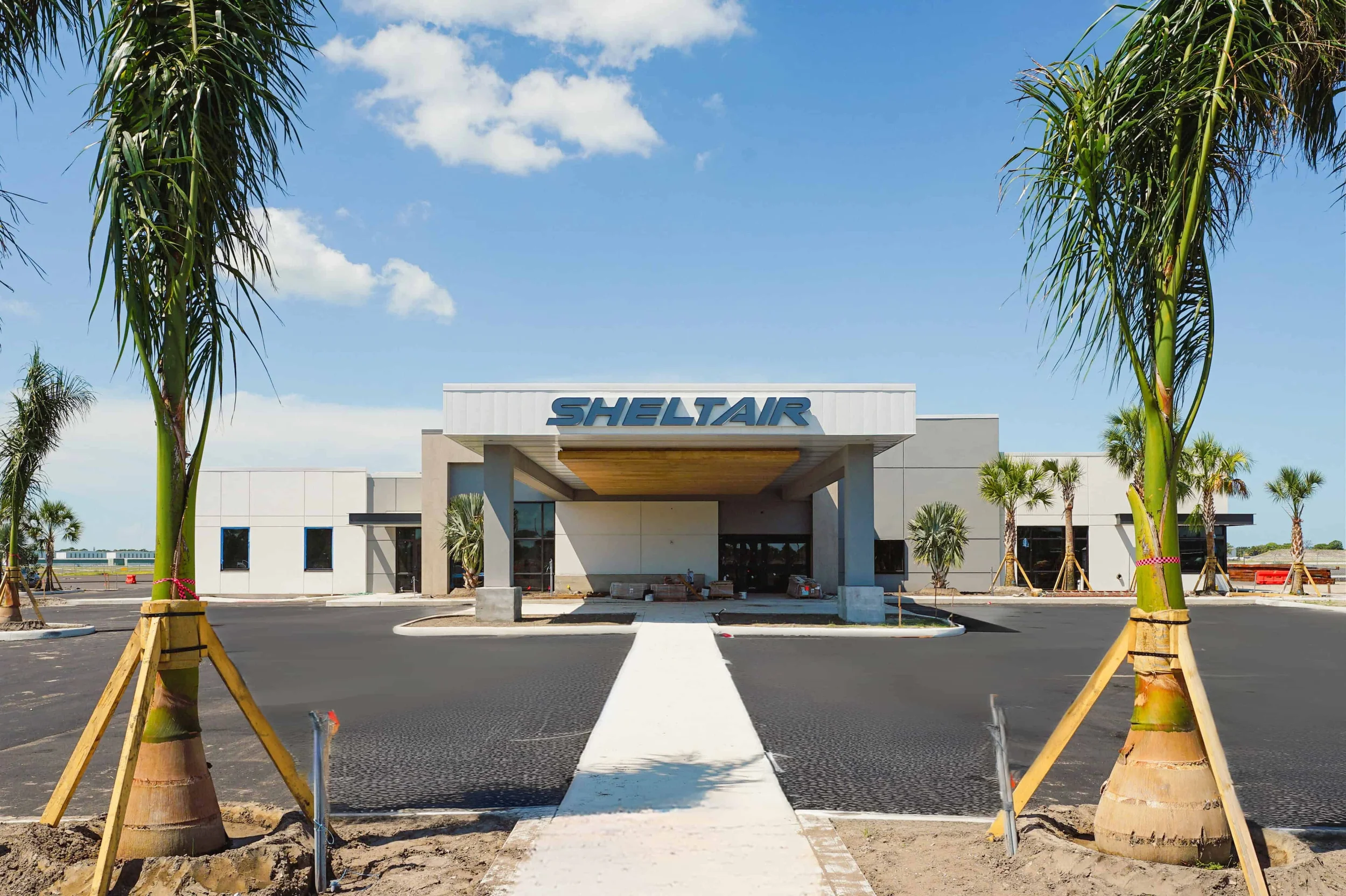 A newly constructed building with a sign that reads 'SHELTAIR'. The building has a modern design with white exterior walls and large windows. There is a concrete walkway leading to the entrance, surrounded by a parking lot, and palm trees flanking the walkway. The sky is blue with some clouds.