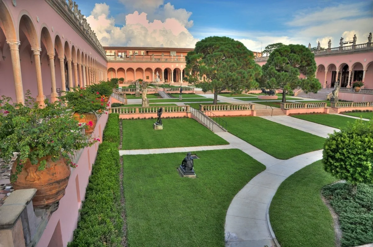Sarasota Motor Club - Sports Car Rental Sarasota FL - A pristine courtyard garden with lush green lawns, statues, and large trimmed trees. Pink Mediterranean-style building with arched walkways surrounds the garden, under a partly cloudy sky.