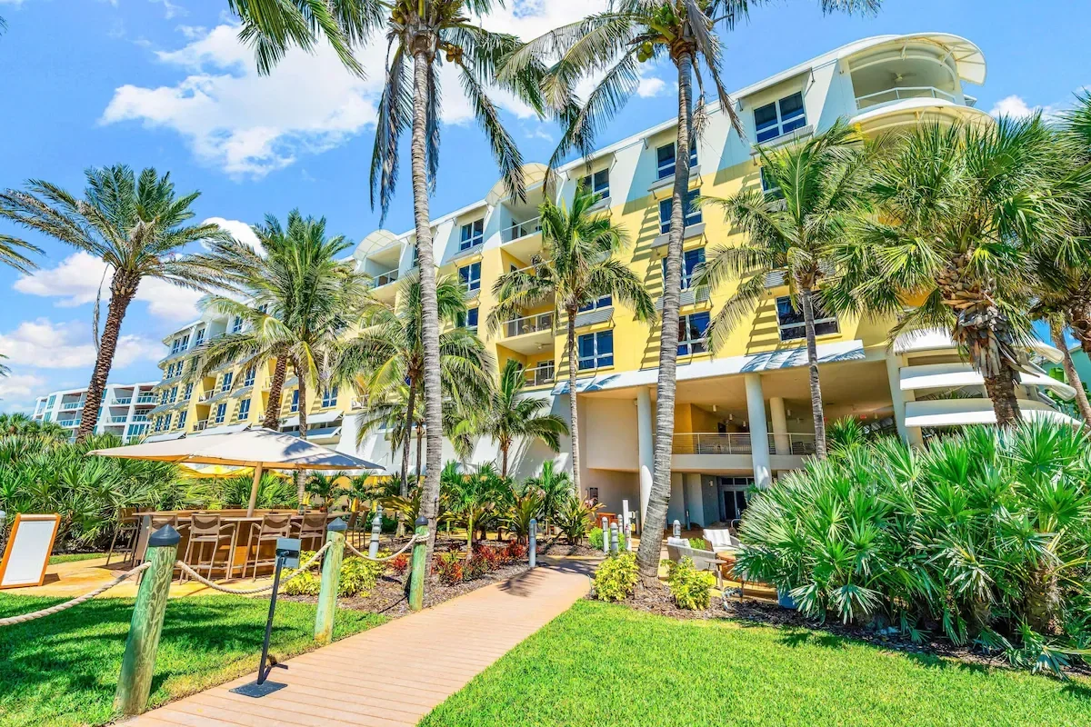 A tropical resort with a yellow building, palm trees, and an outdoor seating area with a large umbrella.