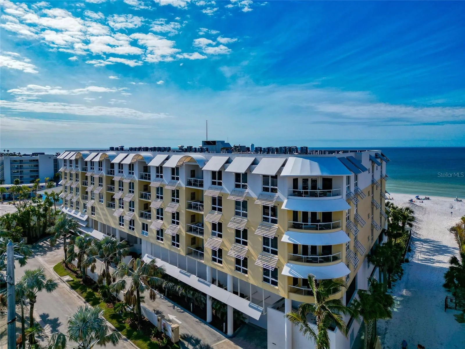 A yellow multi-story beach hotel with white awnings and balconies, palm trees in front, and a sandy beach with people and the ocean in the background under a blue sky with scattered clouds.