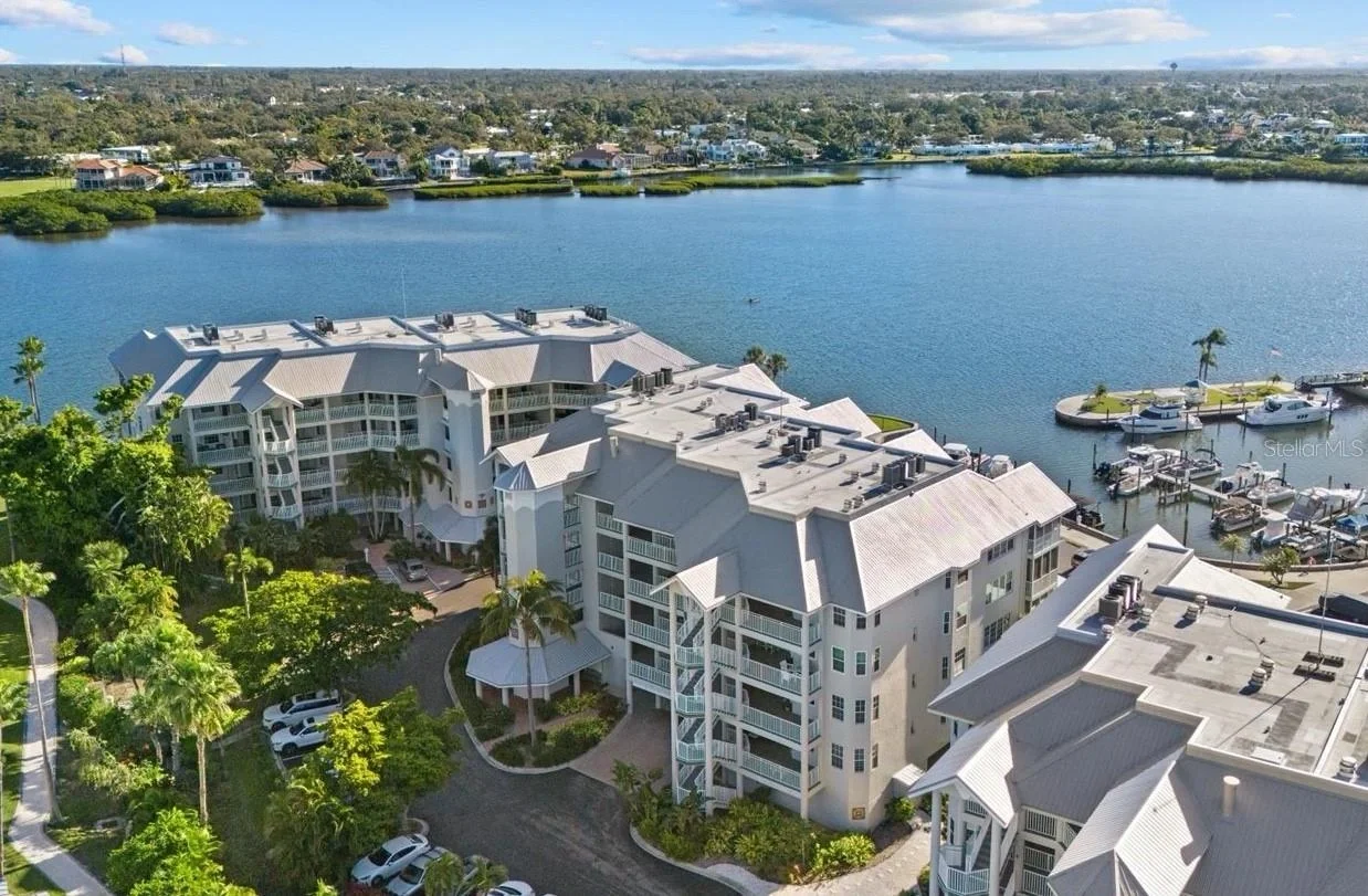 Aerial view of a waterfront apartment complex with white buildings, surrounded by palm trees and lush greenery, overlooking a calm blue body of water with docks and boats.