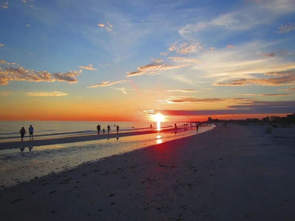 Sunset over the ocean with a colorful sky, people walking along the beach, and reflections on the wet sand.