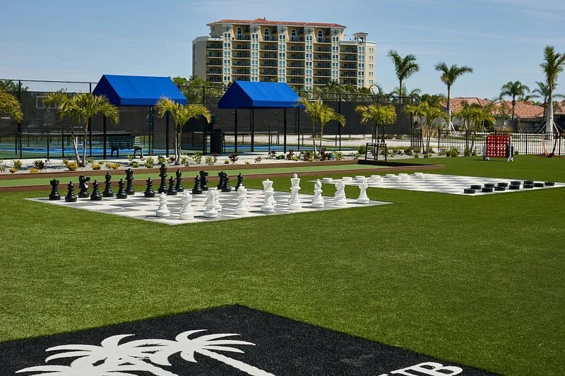 Sarasota Motor Club - Palmetto Marriott luxury car rental - An outdoor chess and checkers play area on a grassy lawn, with palm trees and blue shaded structures, a tennis court.