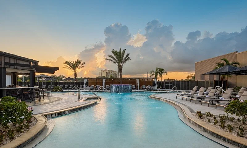 Sarasota Motor Club - Palmetto Marriott luxury car rental - Empty swimming pool at sunset with lounge chairs, umbrellas, palm trees, and cloudy sky.