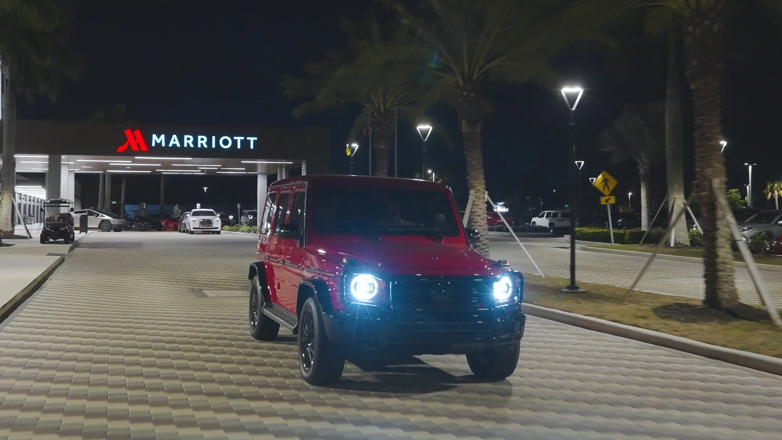 Sarasota Luxury Car Rentals - Palmetto Marriott car rental - Red Mercedes-Benz G-Class SUV parked in front of a Marriott hotel at night, with bright headlights on, palm trees, and other parked cars in the background.