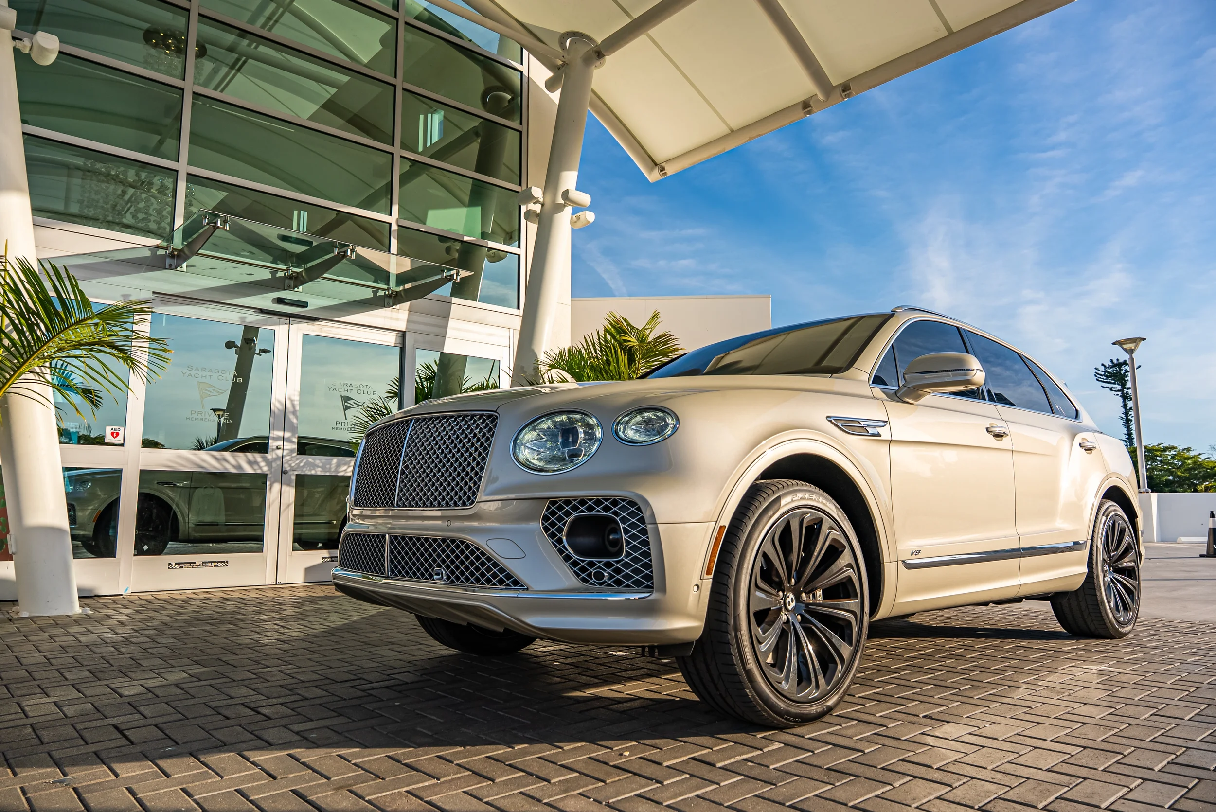 Sarasota Motor Club - Ritz Carlton exotic car rental- A luxury beige SUV parked outside a modern building with glass windows and palm trees, under a blue sky.