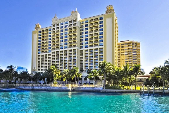 Sarasota Motor Club - Ritz Carlton exotic car rental - A tropical hotel pool area with palm trees, lounge chairs with umbrellas, and cabanas. People are swimming and relaxing on chairs.