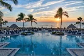 Sarasota Motor Club - Ritz Carlton exotic car rental - A tropical hotel pool area with palm trees, lounge chairs with umbrellas, and cabanas. People are swimming and relaxing on chairs.