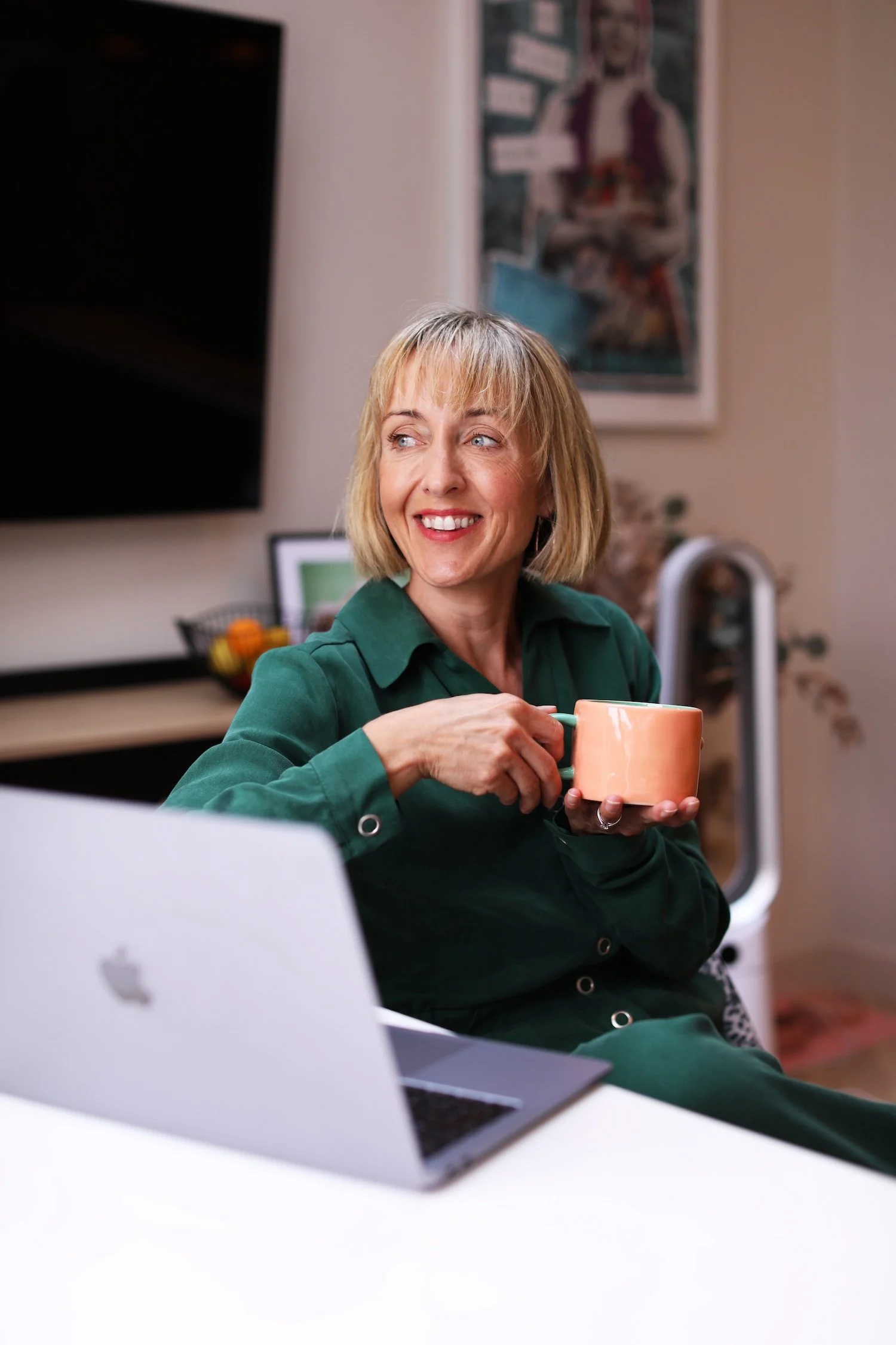 A woman with blonde, shoulder-length hair and blue eyes smiling and holding an orange mug, sitting with a laptop in front of her