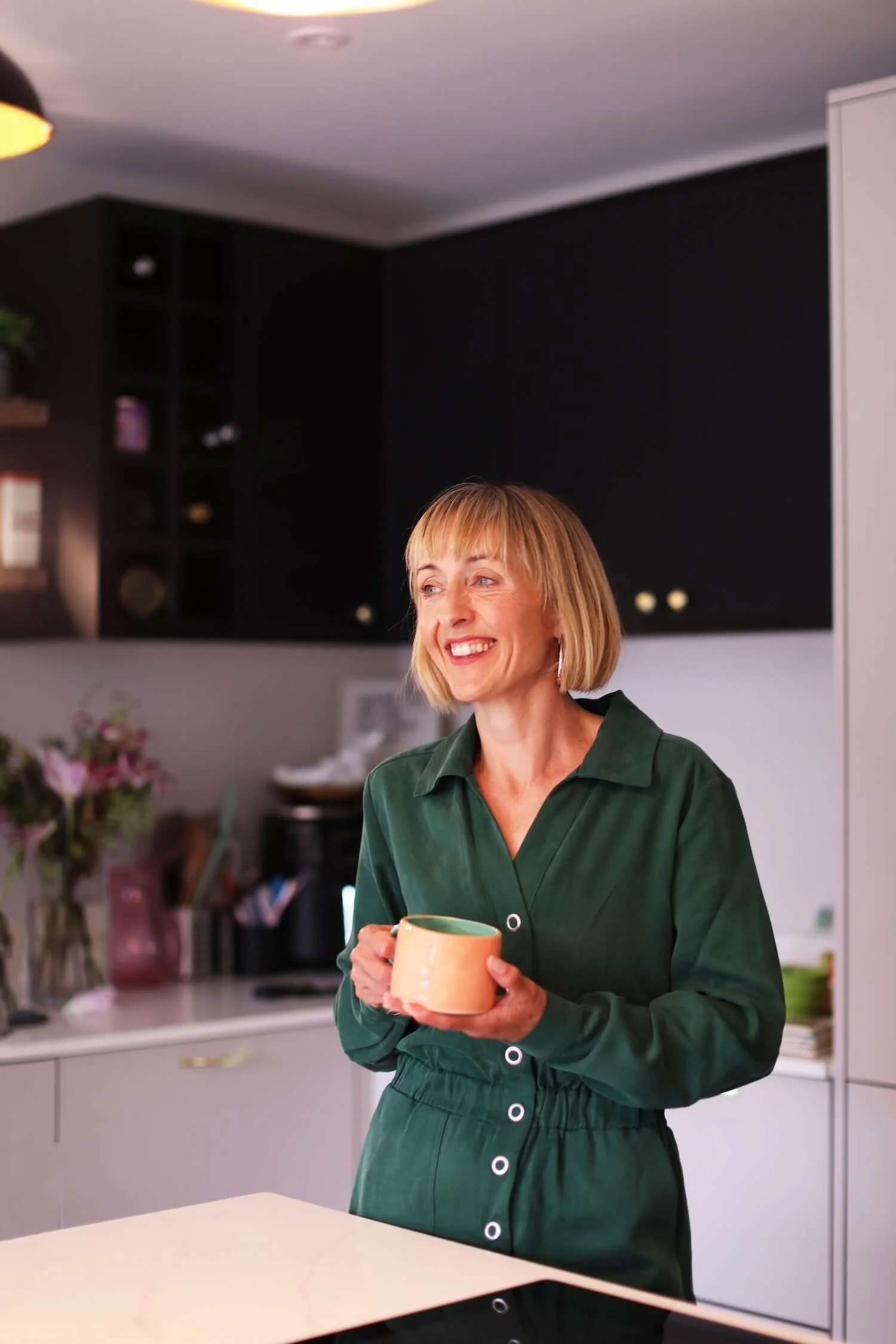 A woman with blond hair wearing a green dress holding a coffee mug in a kitchen