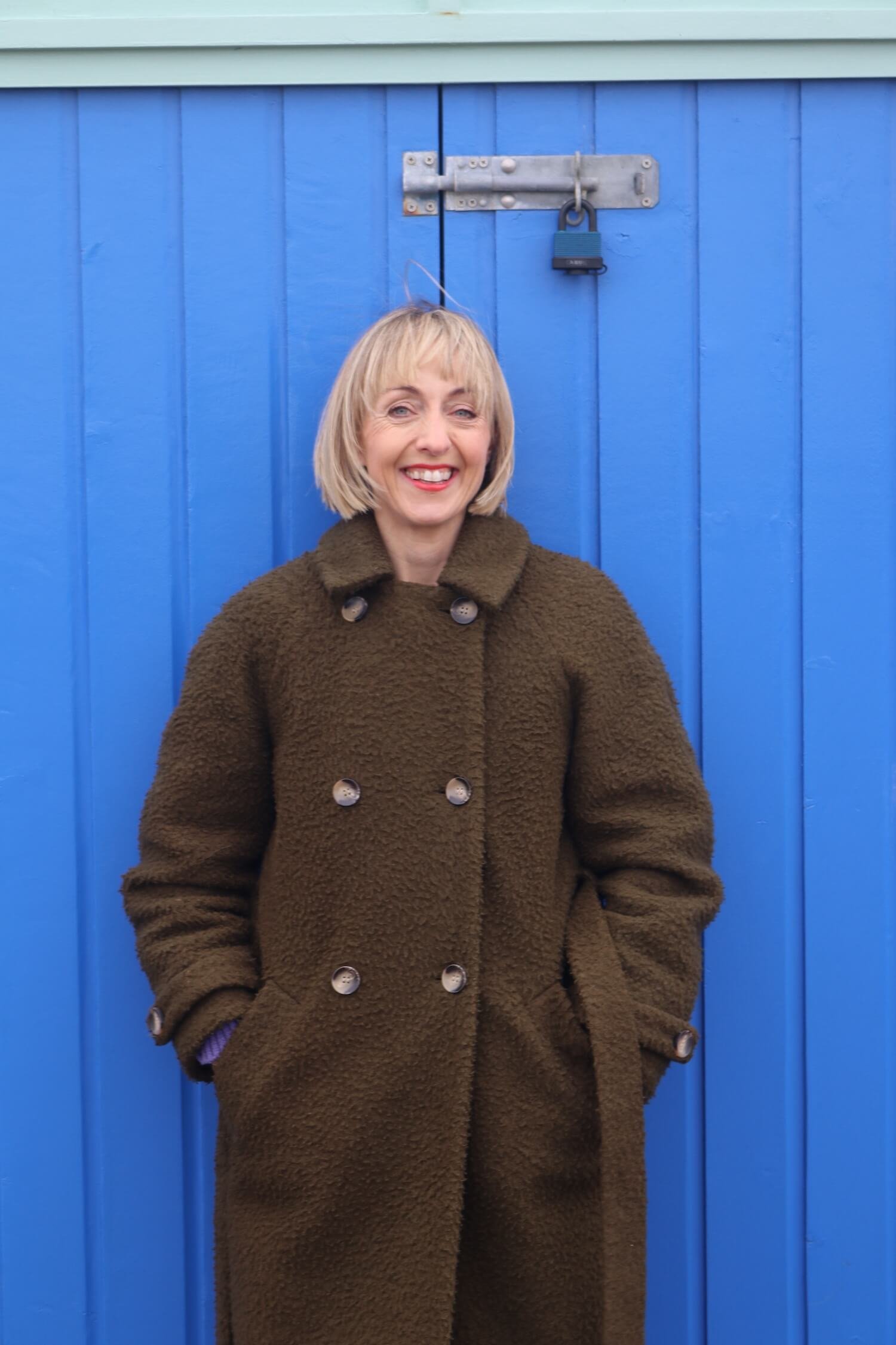 A woman with blonde hair wearing a brown coat with large buttons, standing in front of a blue wooden door with a metal latch and a padlock, smiling at the camera.