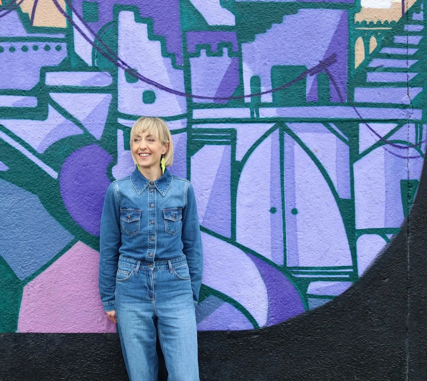 A woman with blonde hair and a denim outfit standing in front of a colorful graffiti wall.