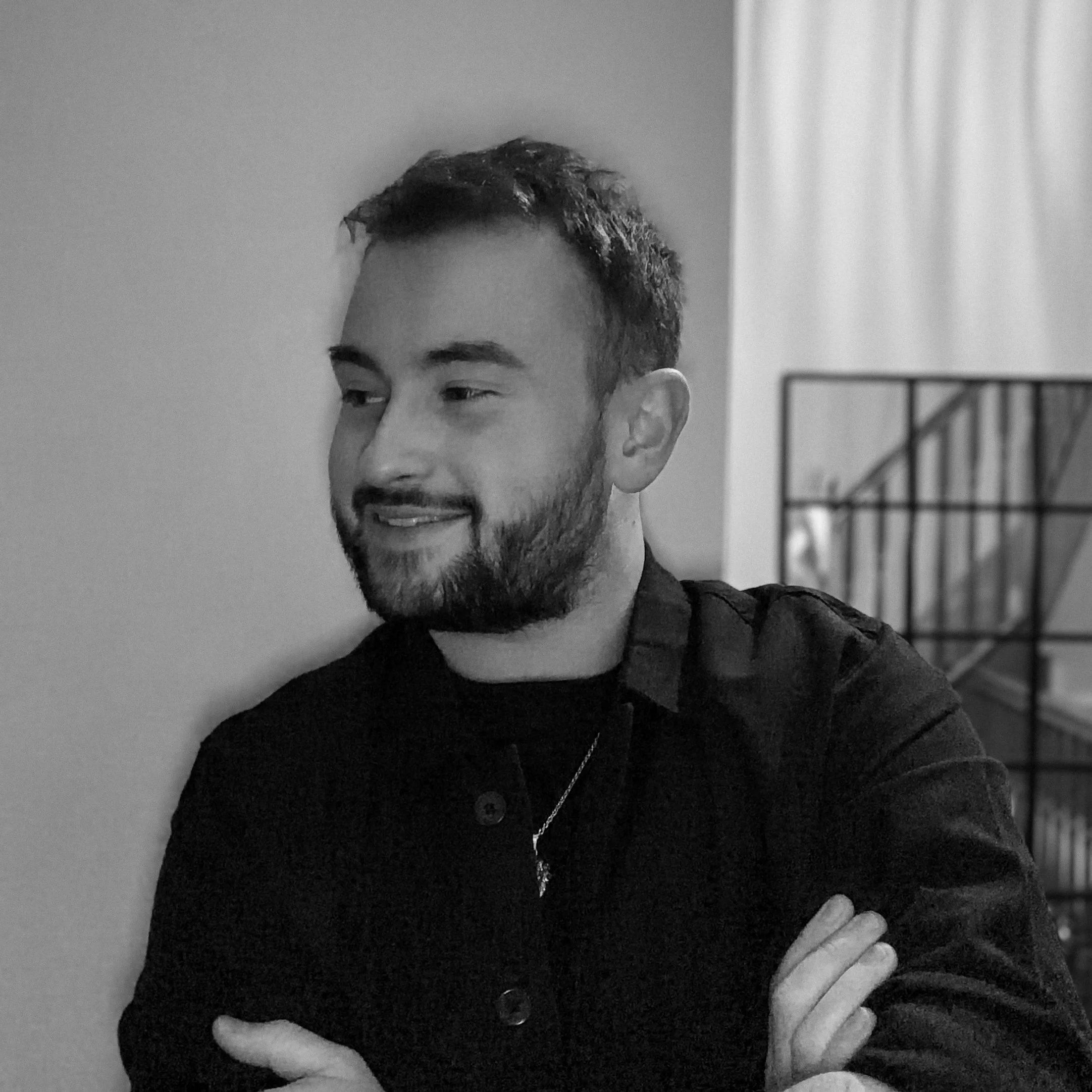 Black and white photo of a man with short hair and facial hair, wearing a dark shirt with buttons. He is smiling slightly and has one hand resting on his chest while the other is crossed over his body. The background shows a blurry interior space with a doorframe or window frame visible.