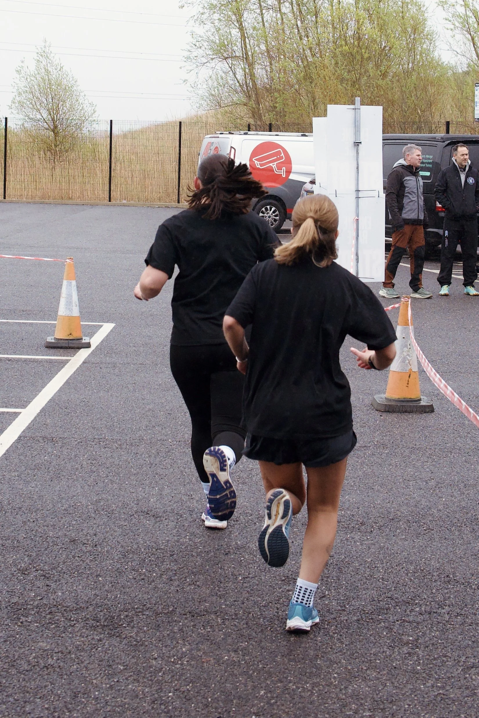 People running outdoors in a parking lot, with cones and a fenced area in the background.