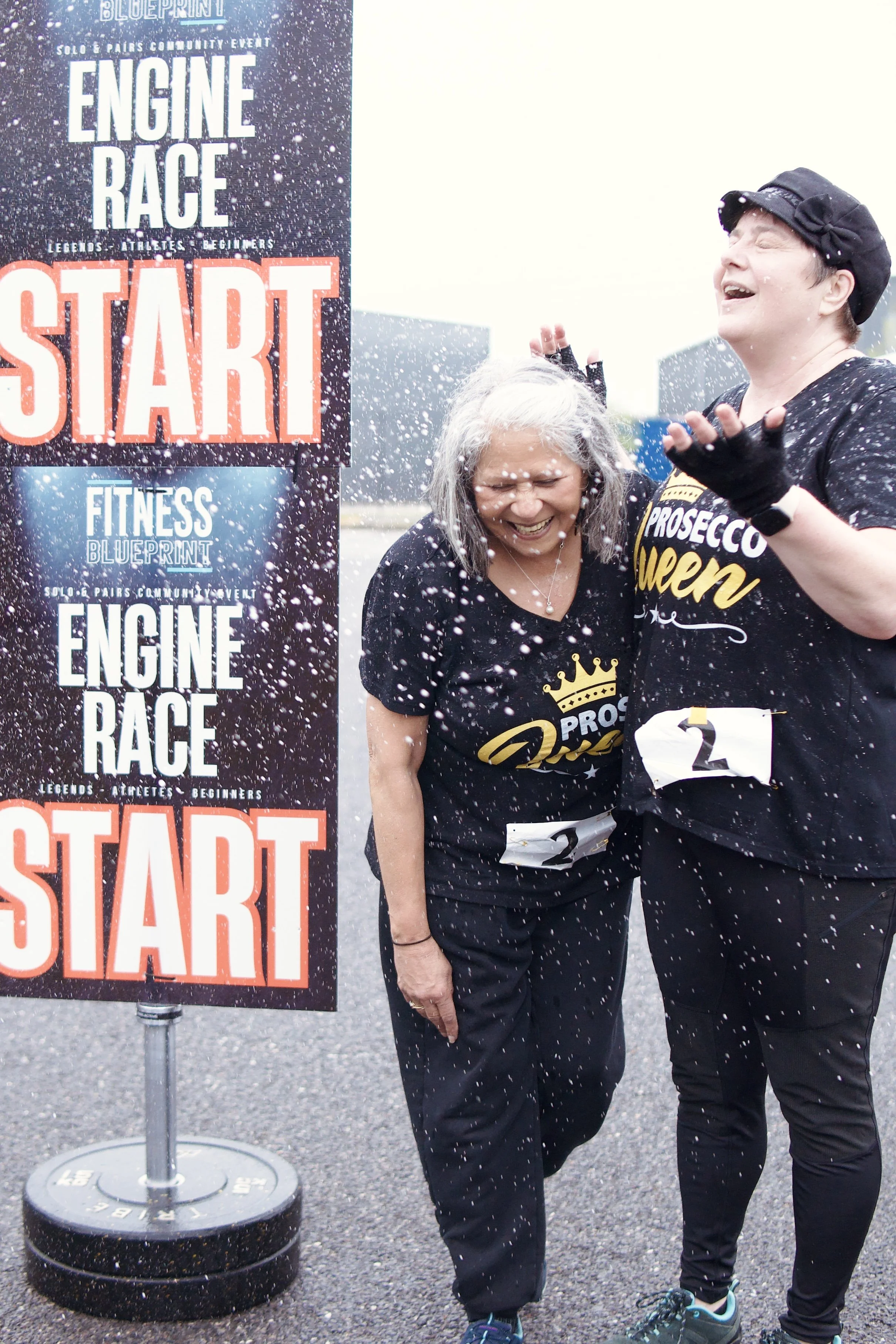 Two women celebrating at the start of a fitness event, standing beside a sign that says 'engine race'. One woman has gray hair and is laughing, while the other woman has short hair and is smiling with her eyes closed. There is confetti or snow falling around them.