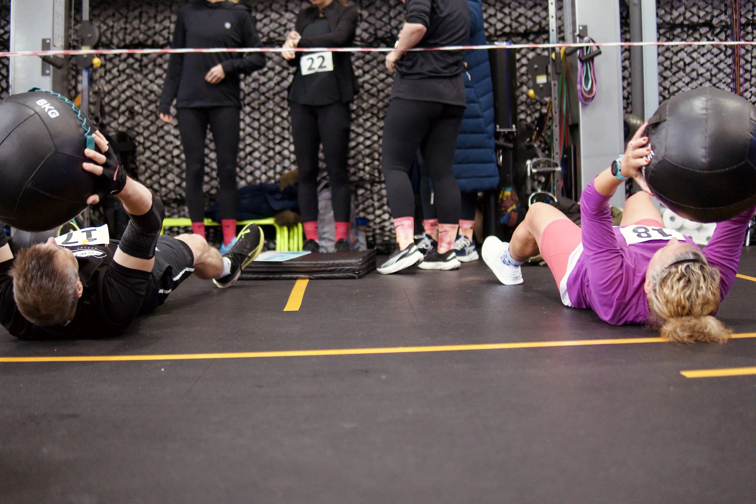 Two people lying on the floor doing an abdominal exercise, holding a medicine ball to their heads. Four other individuals are standing in the background, observing or waiting, in a gym setting with workout equipment.