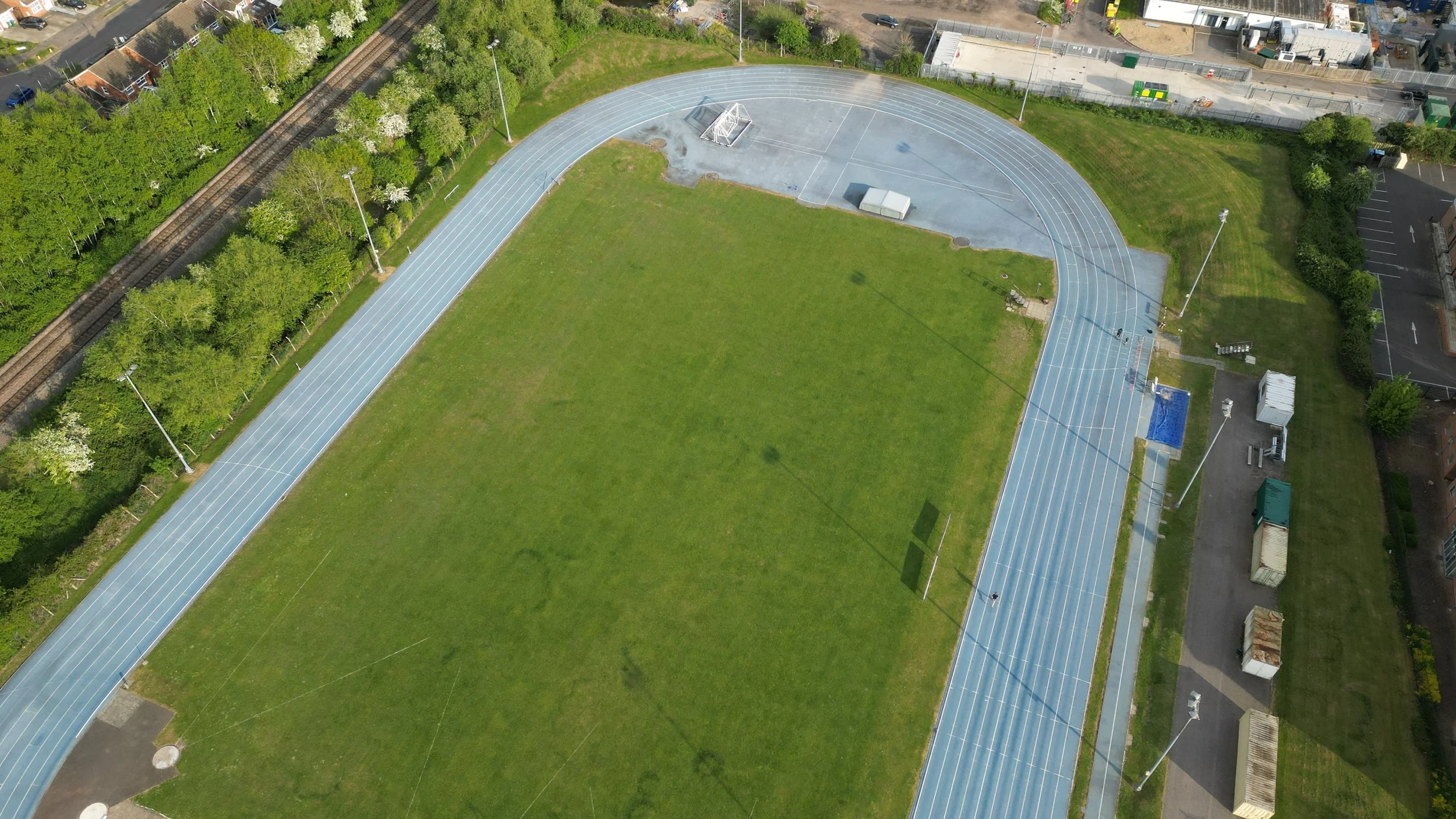 An aerial view of an outdoor running track with lanes, surrounded by greenery, trees, and nearby residential and commercial buildings.