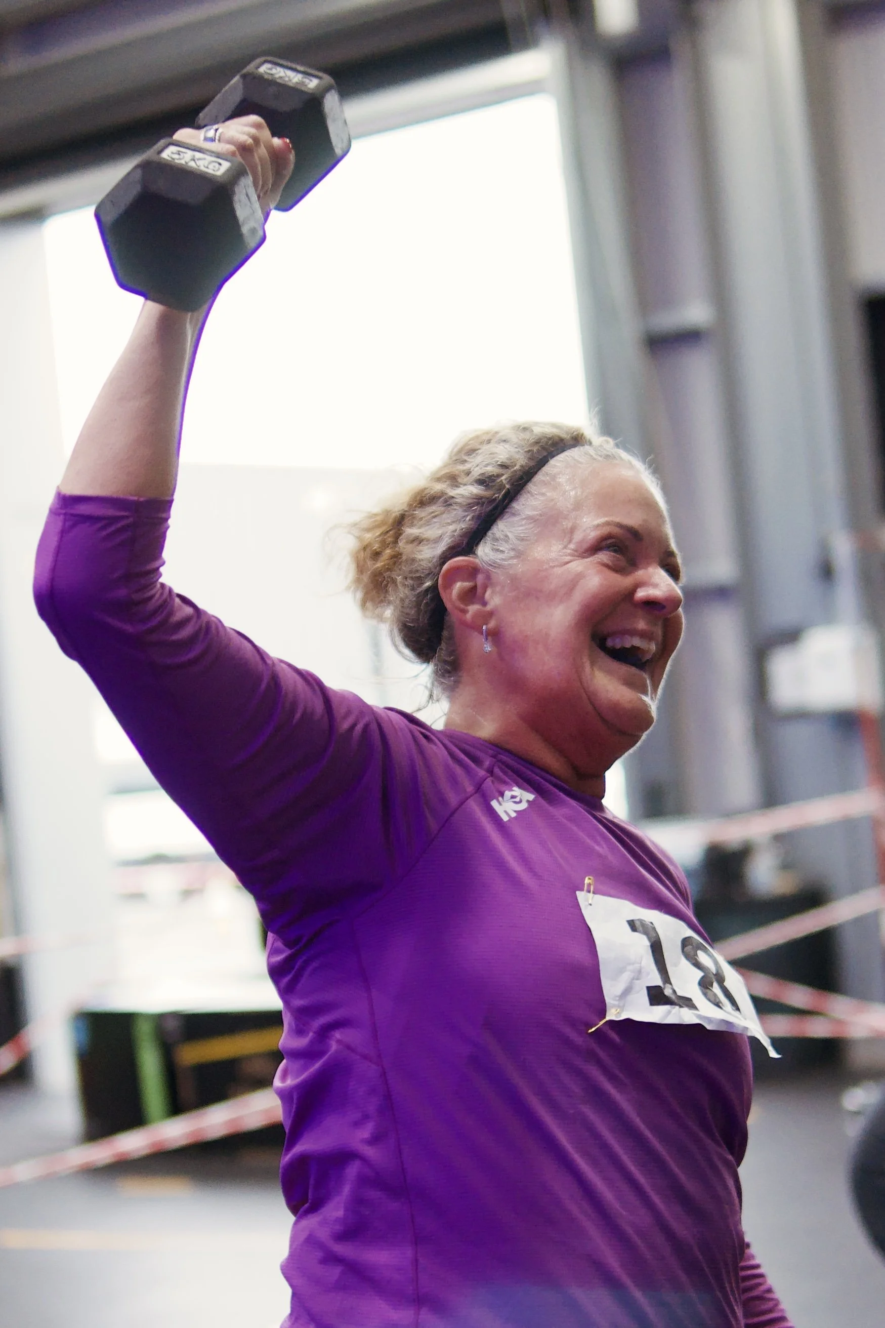 A woman lifting a dumbbell during a workout in a gym, smiling and wearing a purple athletic shirt and a black headband.