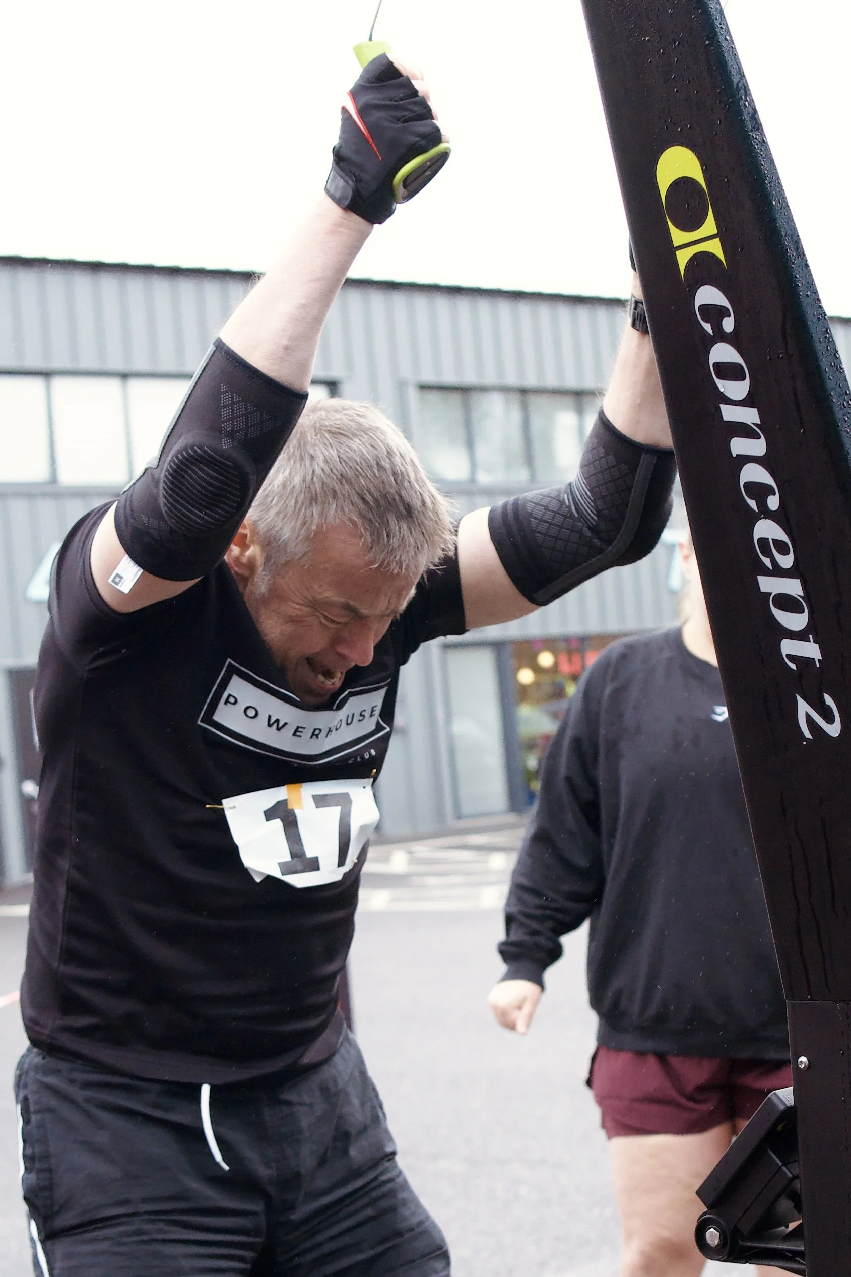 A man with short blonde hair wearing a black athletic shirt with the words 'POWERHOUSE' and the number 17 on it is participating in an outdoor fitness event, raising his arms as if celebrating or stretching. He is wearing black shorts and a black elbow sleeve, with a glove on one hand and a fitness tracker or watch on his wrist. Other participants are visible nearby, and a building with large windows is in the background.