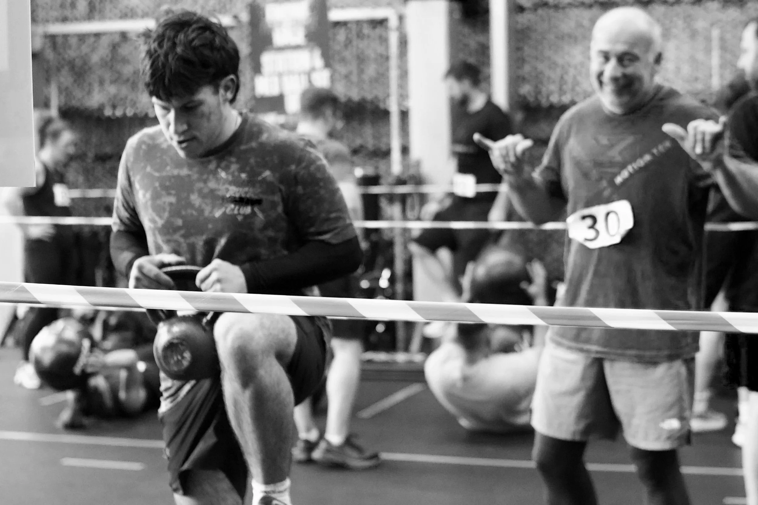 Two men with athlete numbers running in a race inside a sports facility, with blurred spectators and other runners in the background.