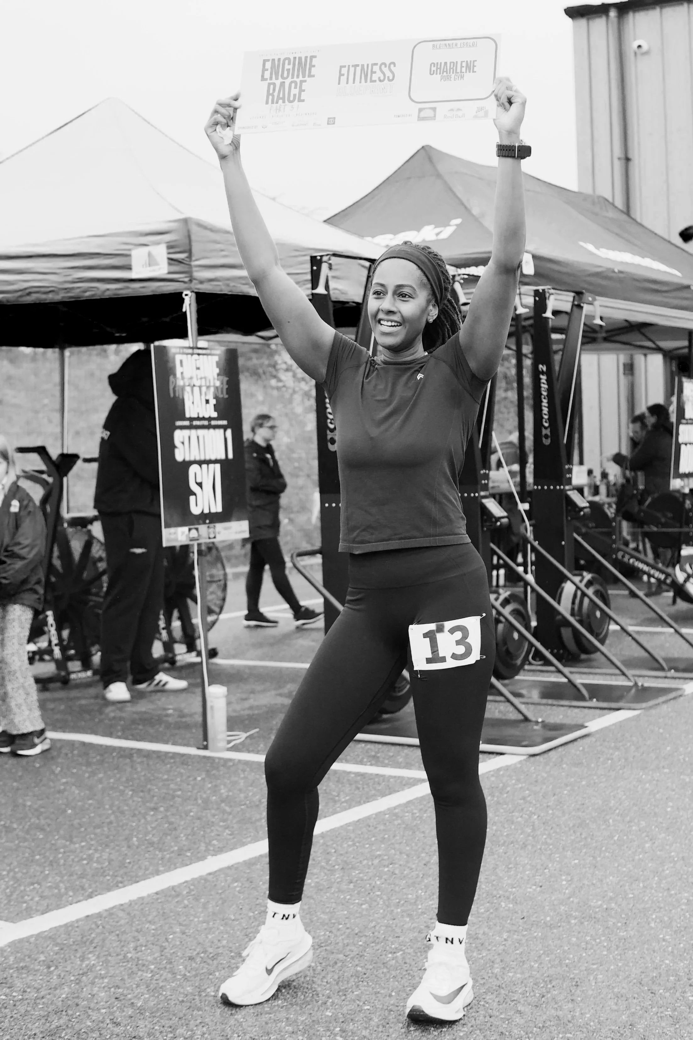 A woman with athletic clothing, a sports watch, and a race bib numbered 13, stands with her arms raised, smiling at a race event. Behind her, there are tents, race signage, and other people preparing or spectating.