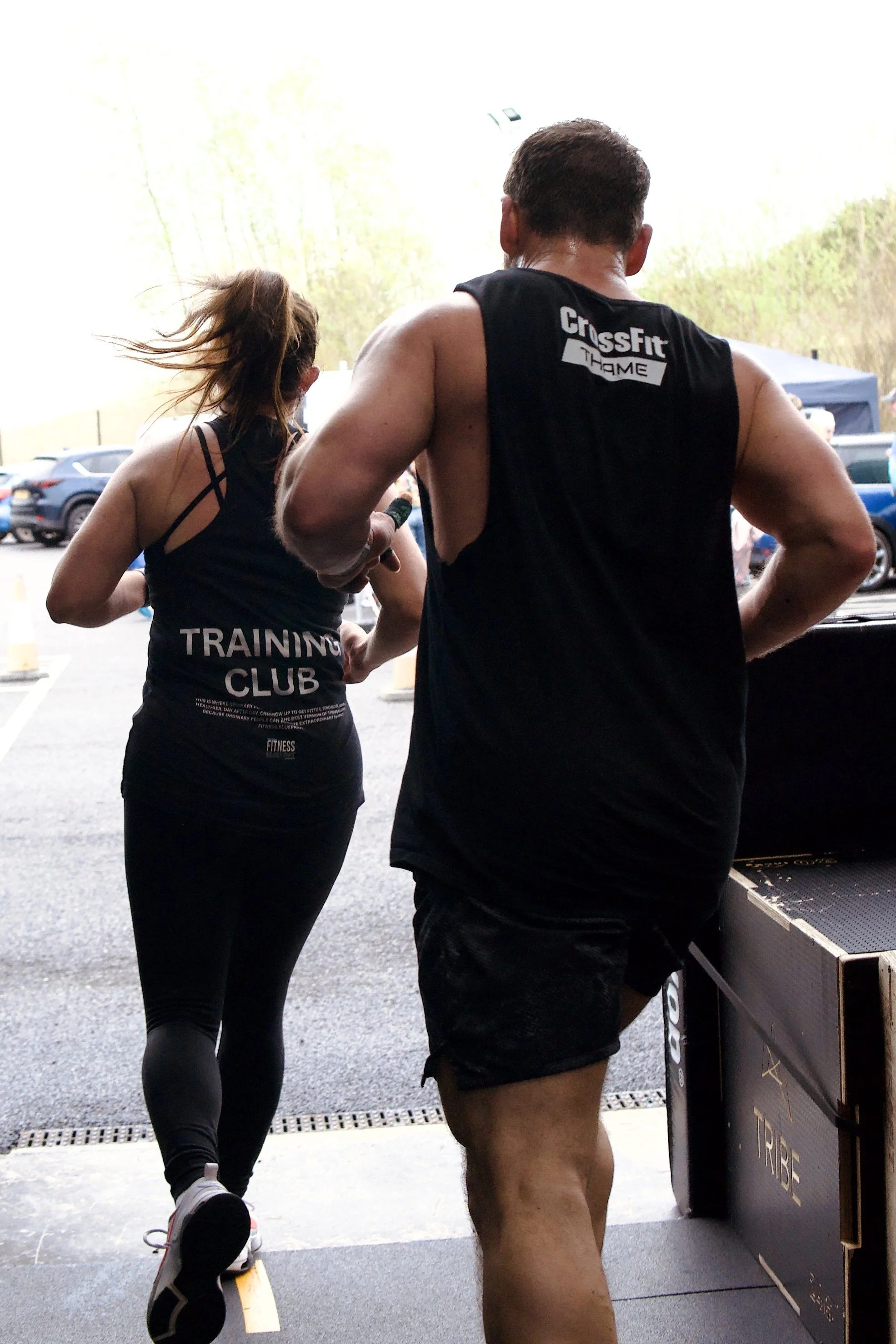 Back view of a man and woman participating in a CrossFit workout, running or jumping over a box outdoors, with cars parked in the background.