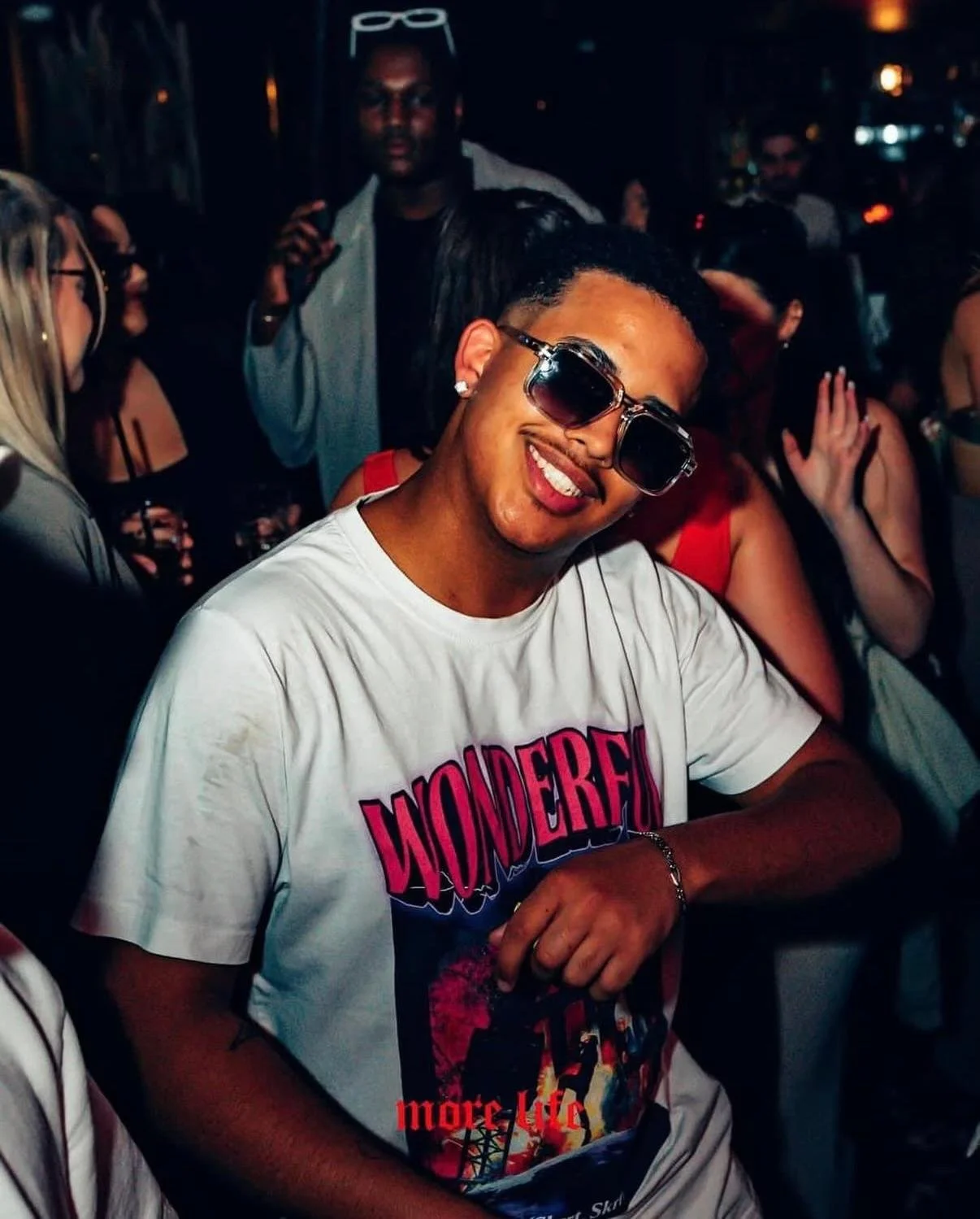 A smiling man wearing sunglasses and a white T-shirt with colorful graphic and the word 'WONDERFUL' on it, at a lively party or social gathering at night.