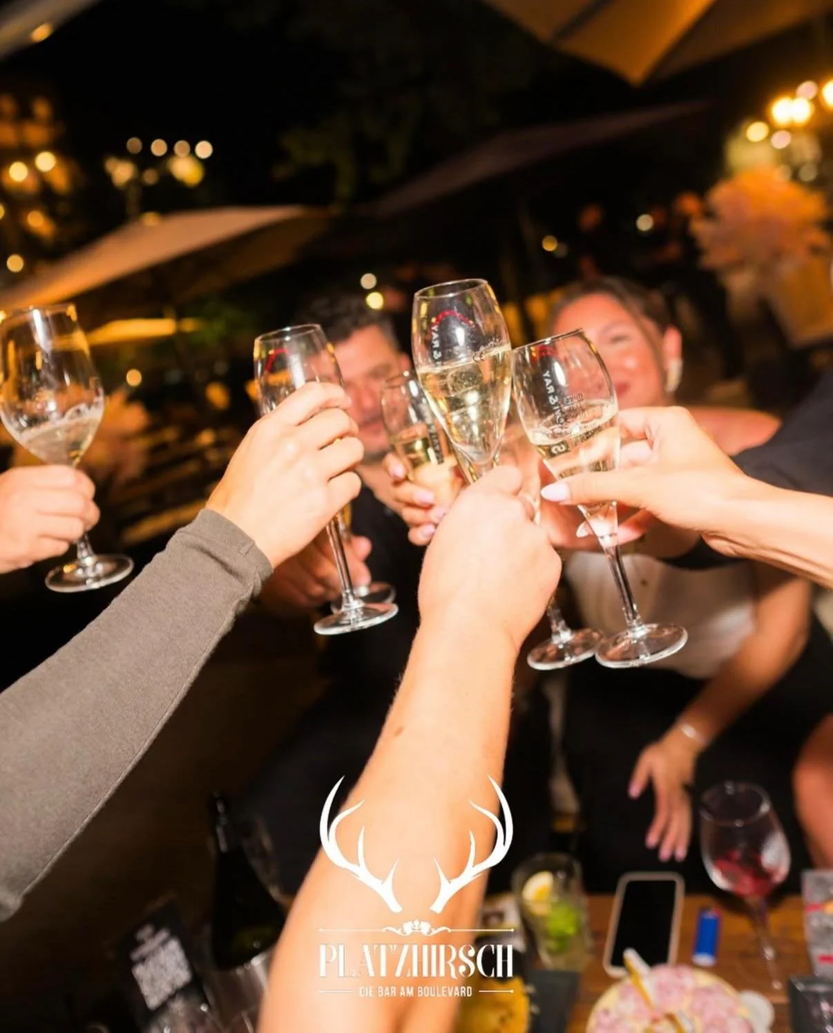 People raising champagne glasses in a toast at a bar or restaurant during night time, with a blurred background and warm lighting.