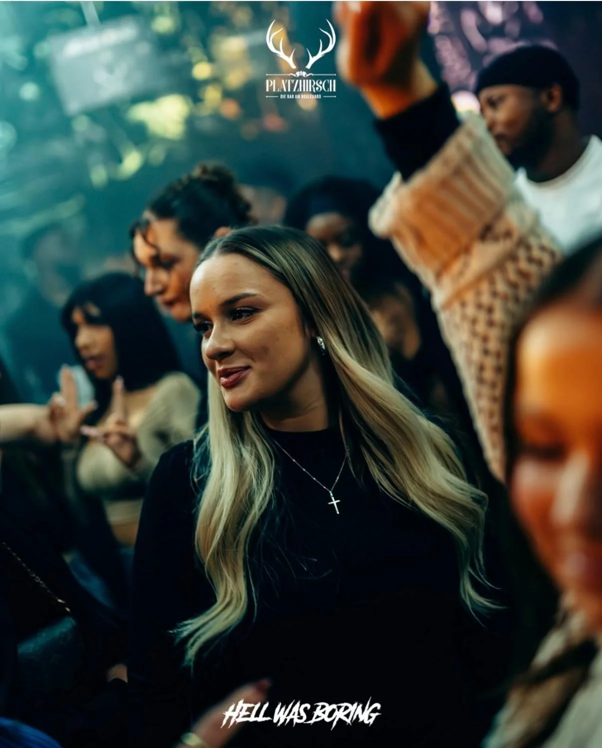 A group of young adults at a social gathering or party, with a woman with long blonde hair and a cross necklace in the foreground. The crowd appears engaged and the setting is lively, with a logo of antlers and text reading 'PLATZHIRSCH' at the top, 