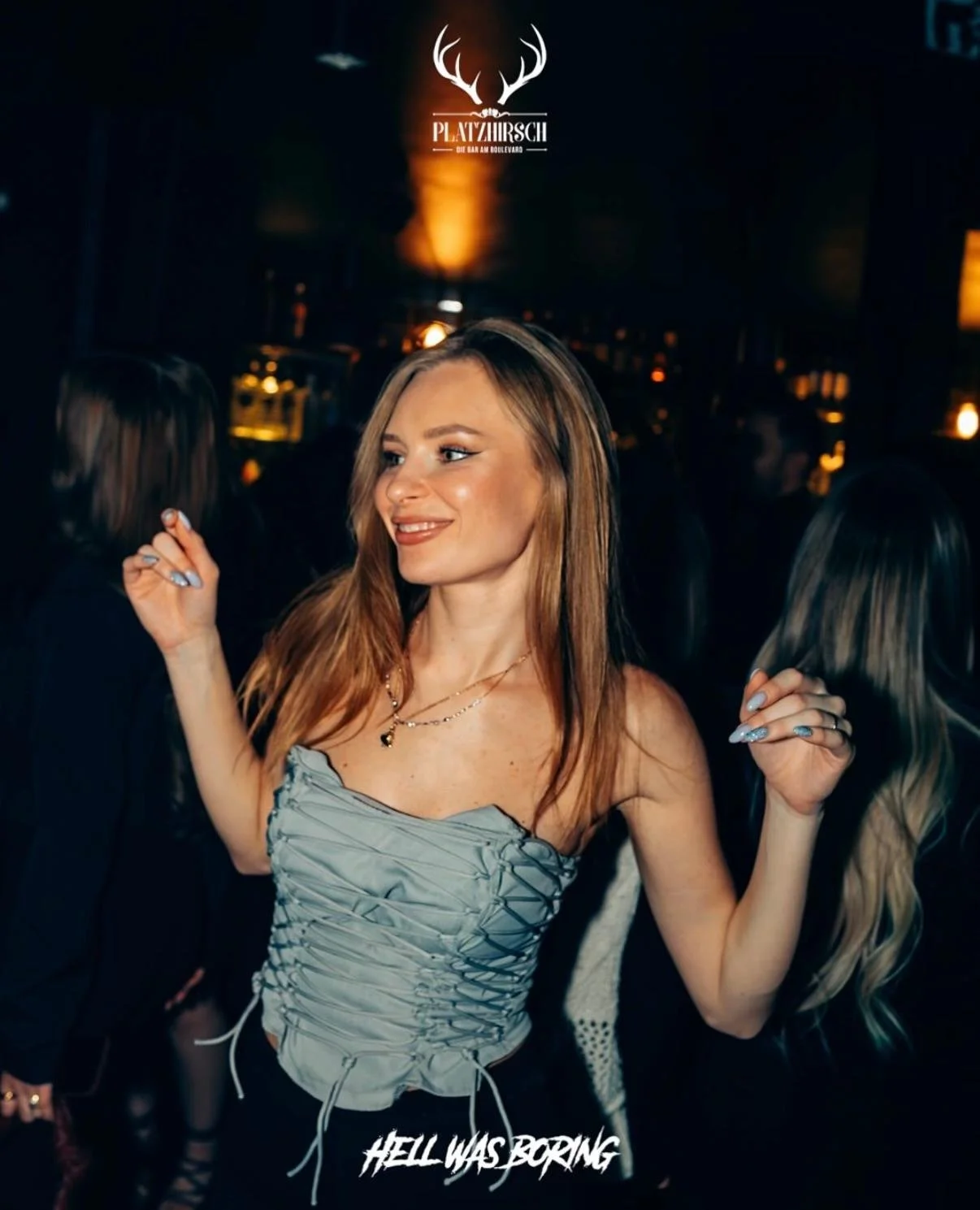 A young woman with long red hair dancing and smiling in a dark, lively club or bar with the text 'Hell Was Boring' at the bottom and a logo with antlers at the top.