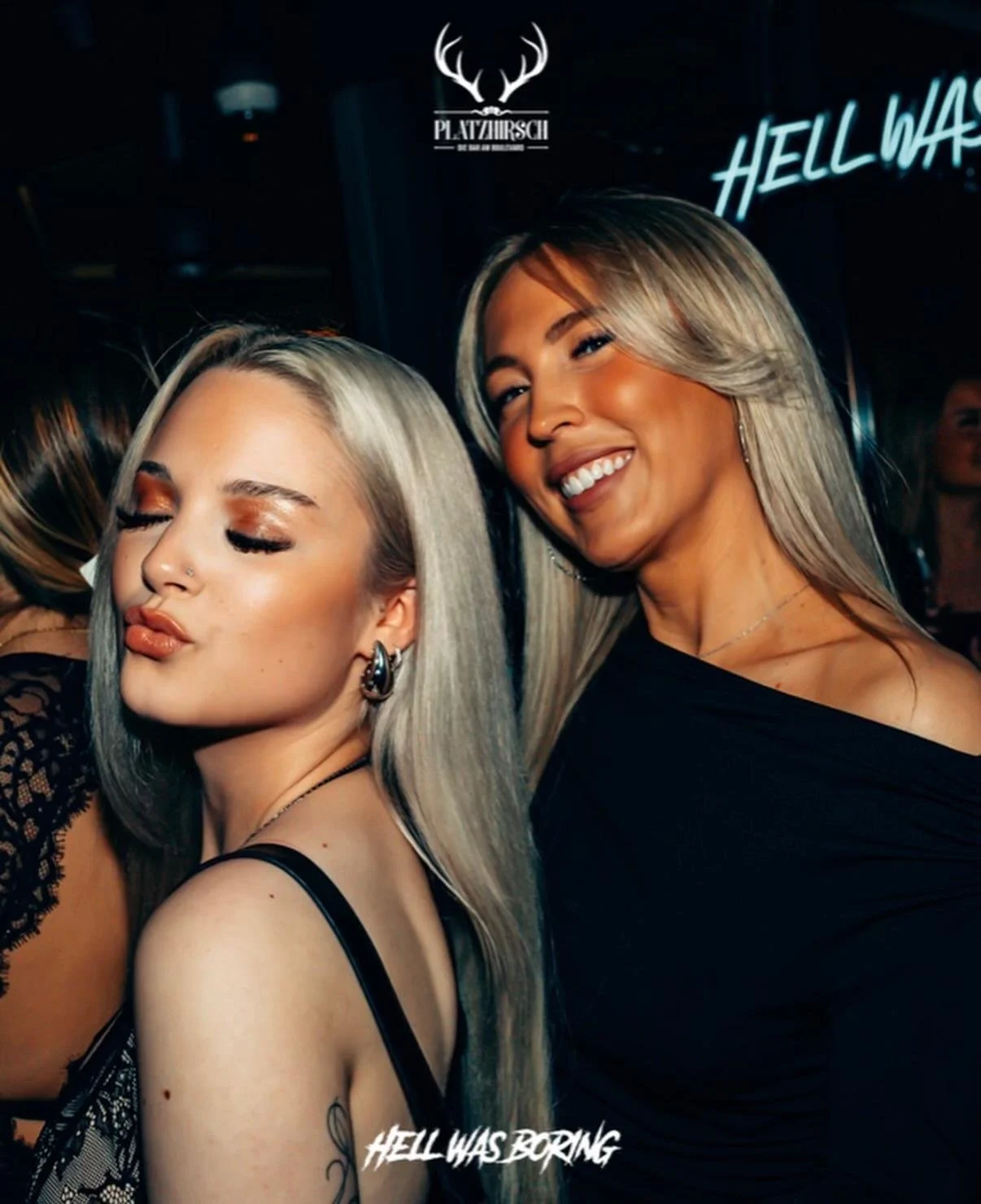 Two women with long hair enjoying themselves at a party. One woman has platinum blonde hair and is wearing a black top, while the other has light brown hair and is smiling brightly. The background has neon signs that say "HELL WAS BORN" and "PLATZHIR