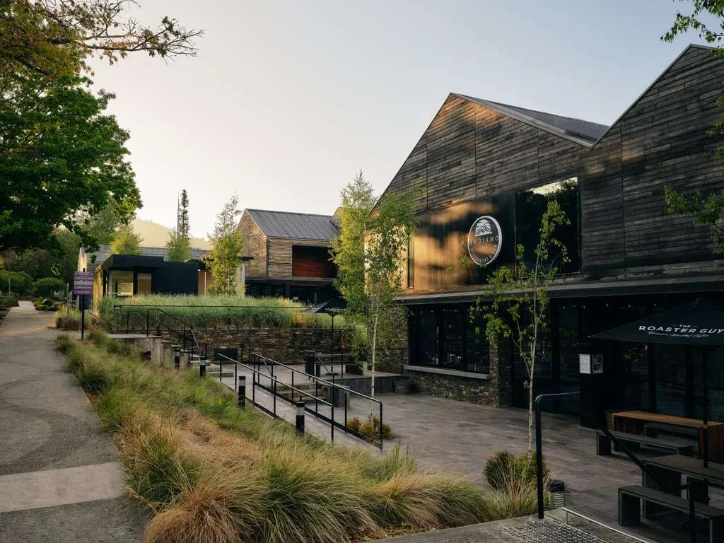 Exterior of a modern building with wooden siding, trees, and outdoor seating, including tables with umbrellas, in a landscaped area with grass and shrubs.