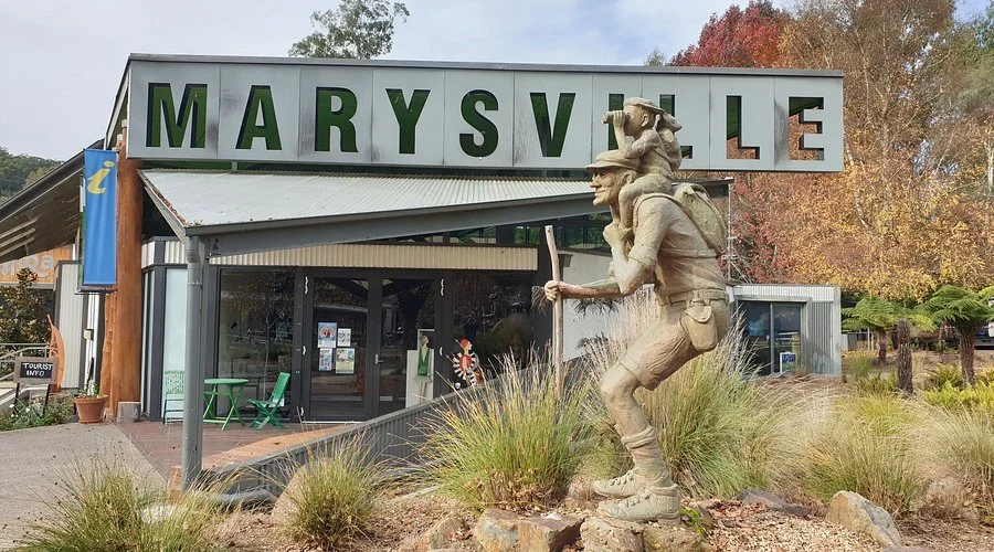A large metallic sign spelling 'MARYSVILLE' with each letter on a separate panel, mounted on a building. In front of the building is a sculpture of a person with a backpack and hat, standing and looking through binoculars. There are plants and trees around, indicating an outdoor setting.