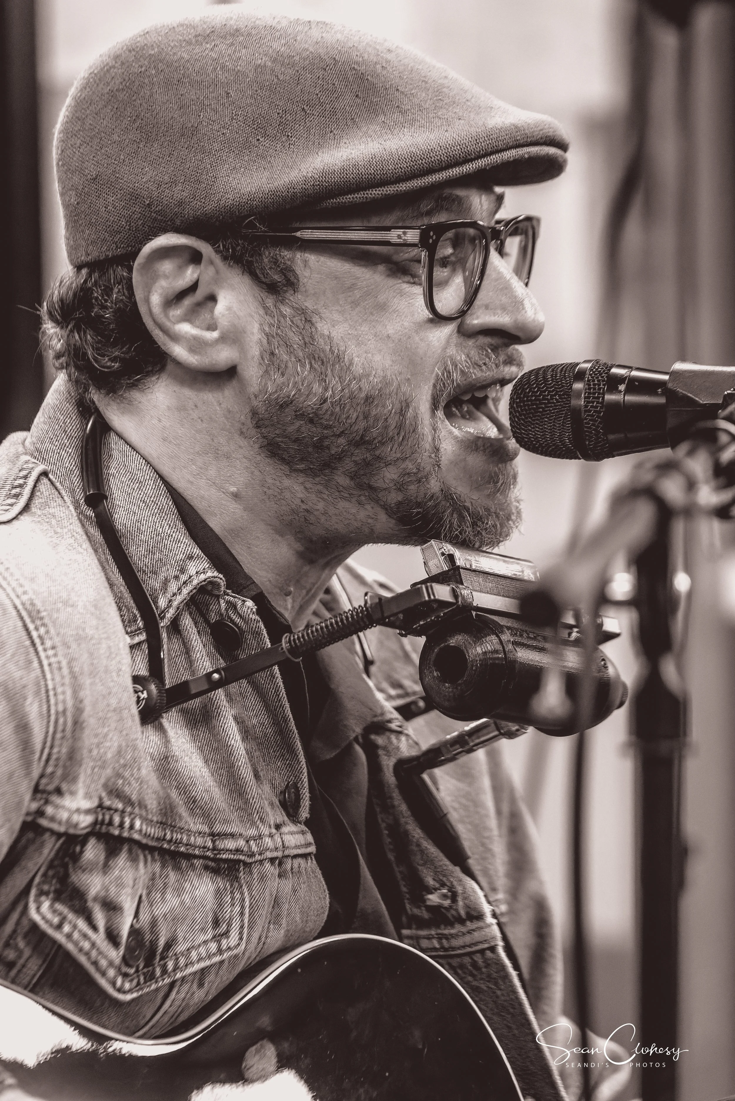 Black and white photo of a man singing into a microphone while playing an acoustic guitar. He is wearing glasses, a fedora-style hat, and a denim jacket.