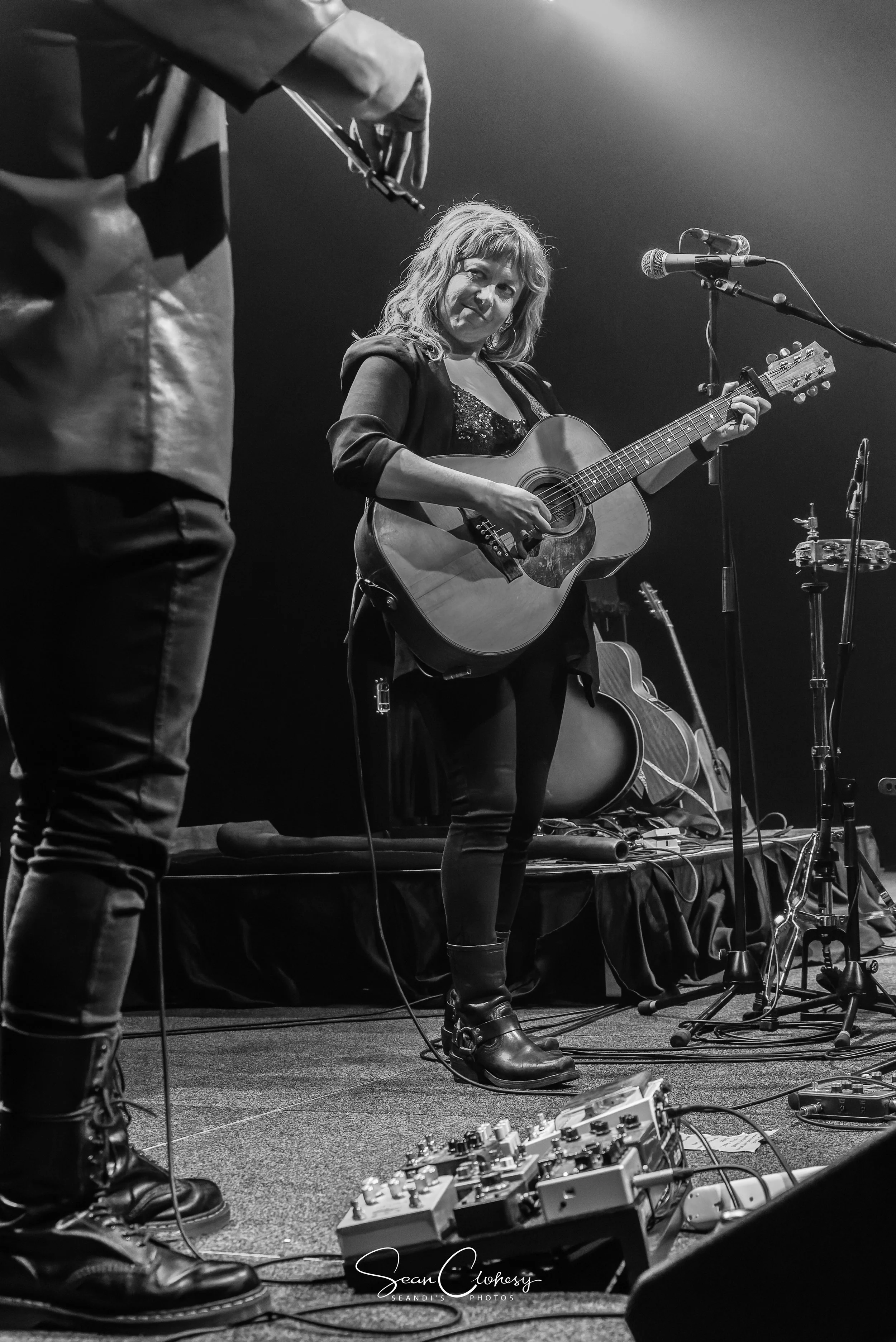 Brooke Taylor smiling while playing an acoustic guitar on stage with microphone stand and pedalboards visible. The image is in black and white.