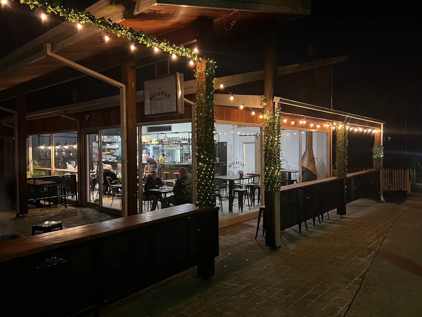 Night scene of a cozy restaurant with glass walls, warm lighting, plants wrapped in string lights, and customers dining outside.