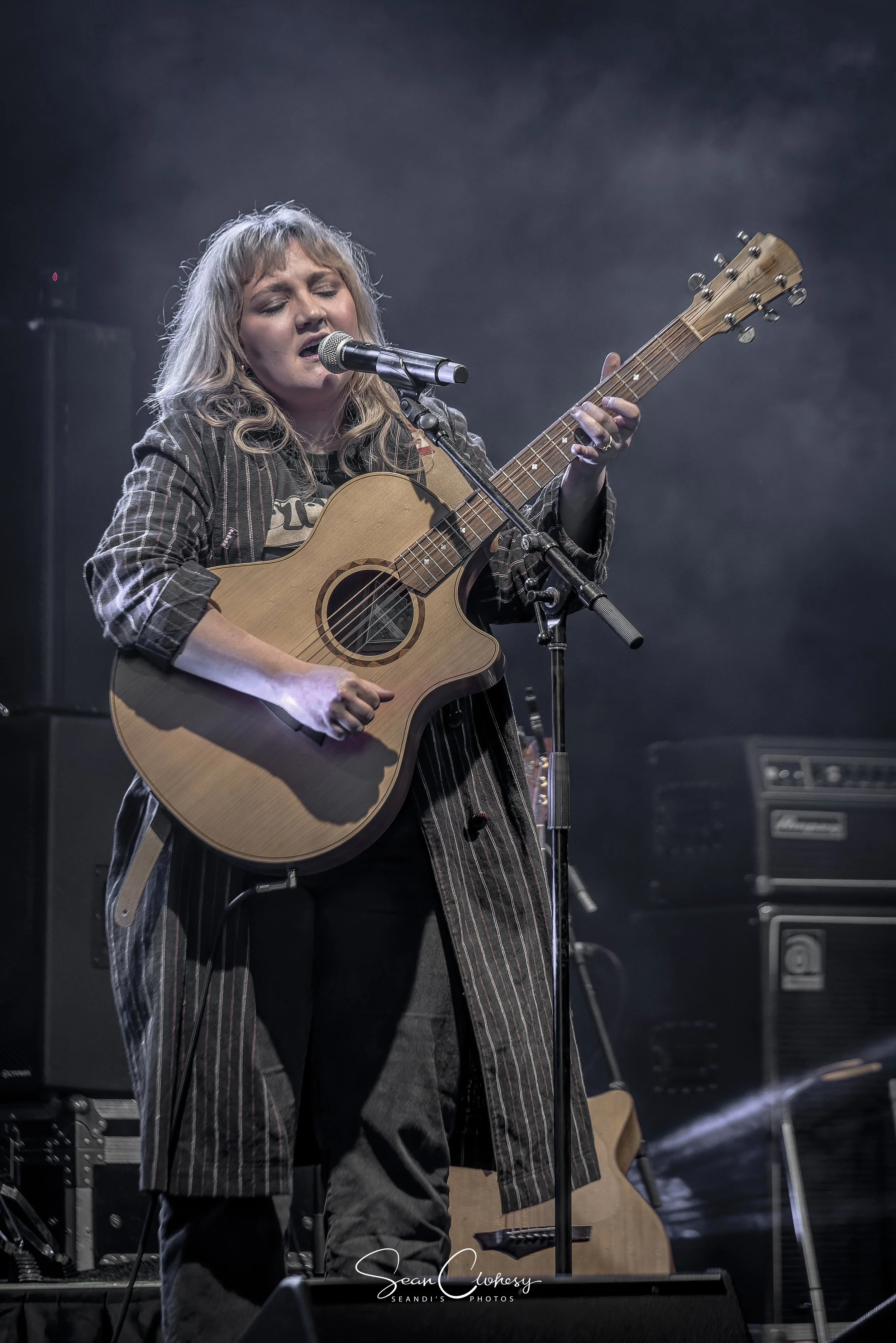 Liv Cartlidge singing into a microphone while playing an acoustic guitar on stage, surrounded by musical equipment.