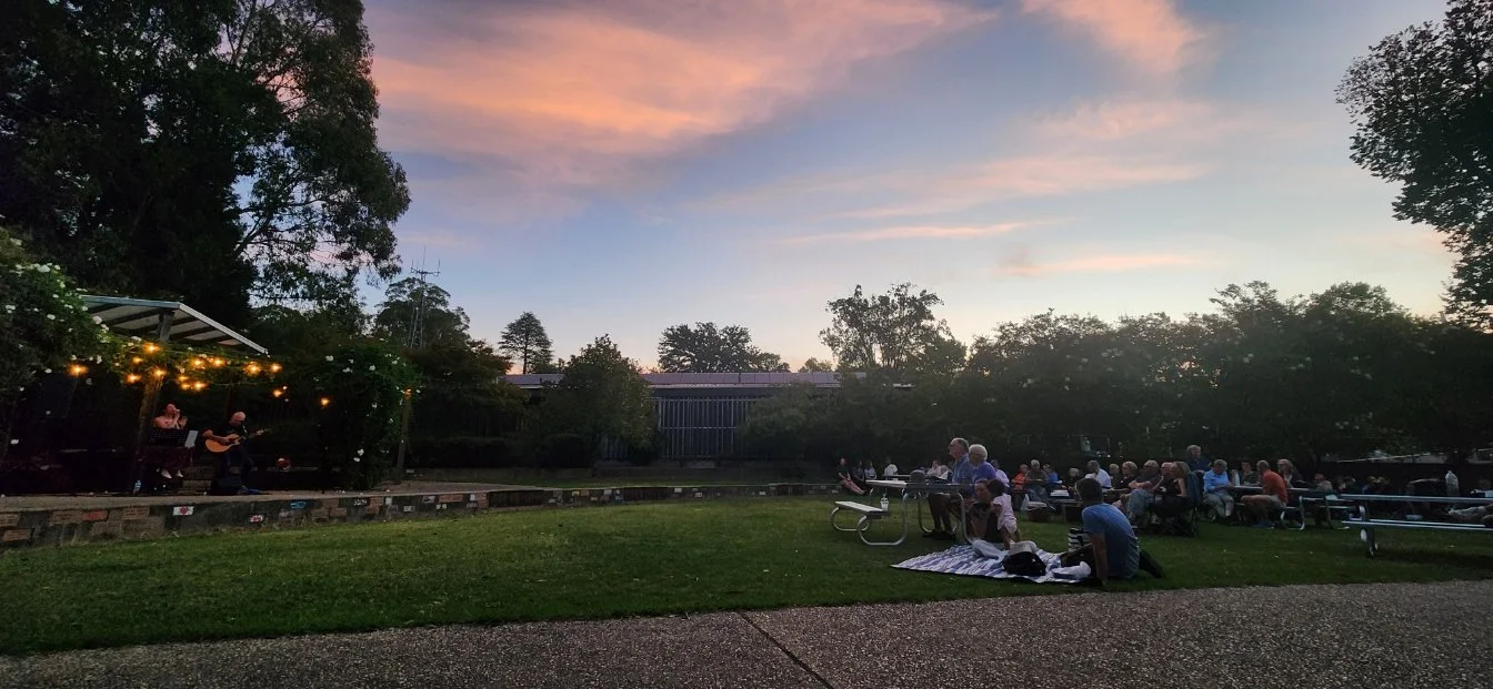 Outdoor concert at sunset with people sitting on picnic blankets and benches, watching a live music performance on stage with string lights, surrounded by trees.