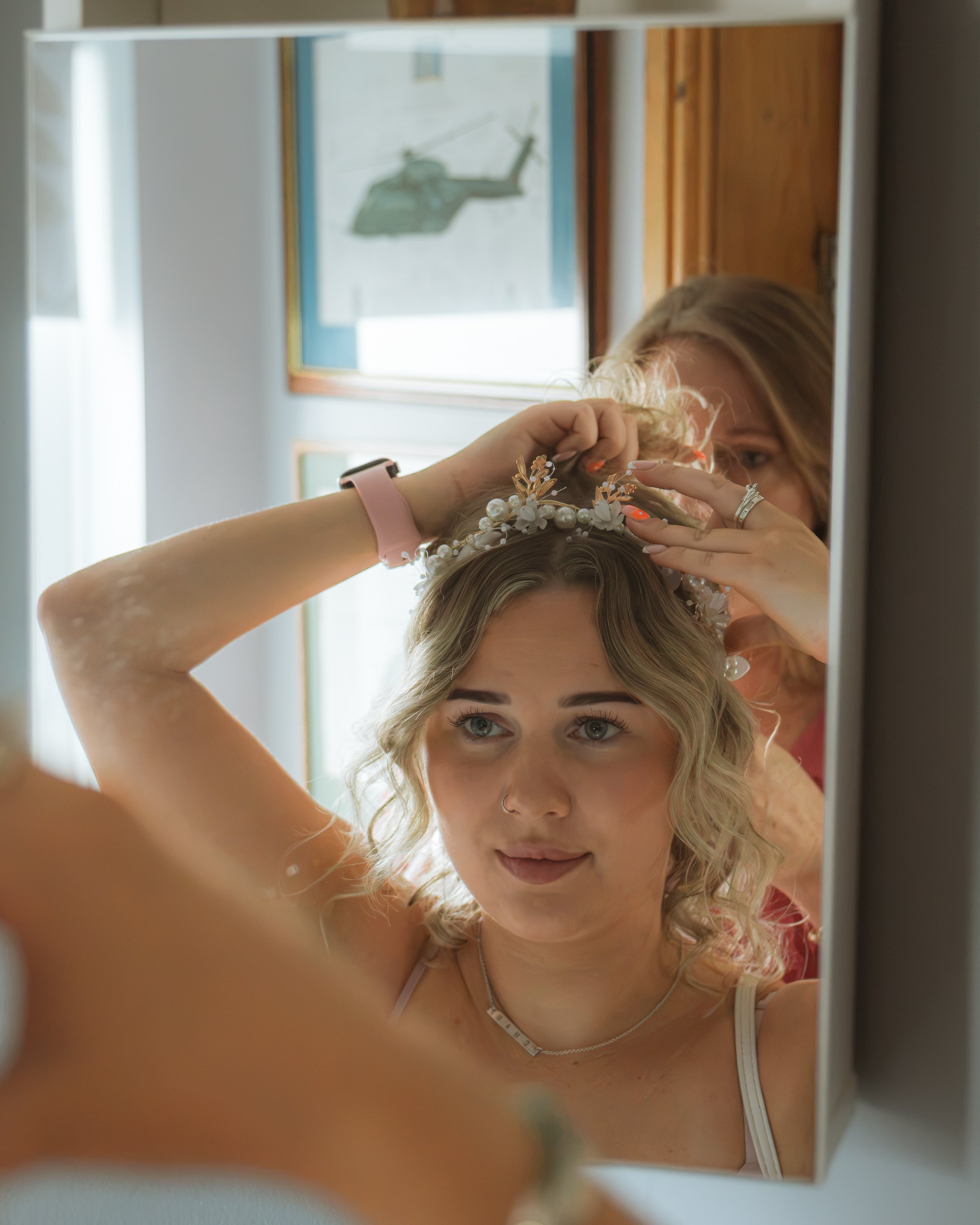 A young woman with blonde wavy hair looking into a mirror, adjusting a decorative floral headpiece. Her face is reflected in the mirror, with her eyes and nose visible. The reflection shows her upper body and the background includes a window, a woode