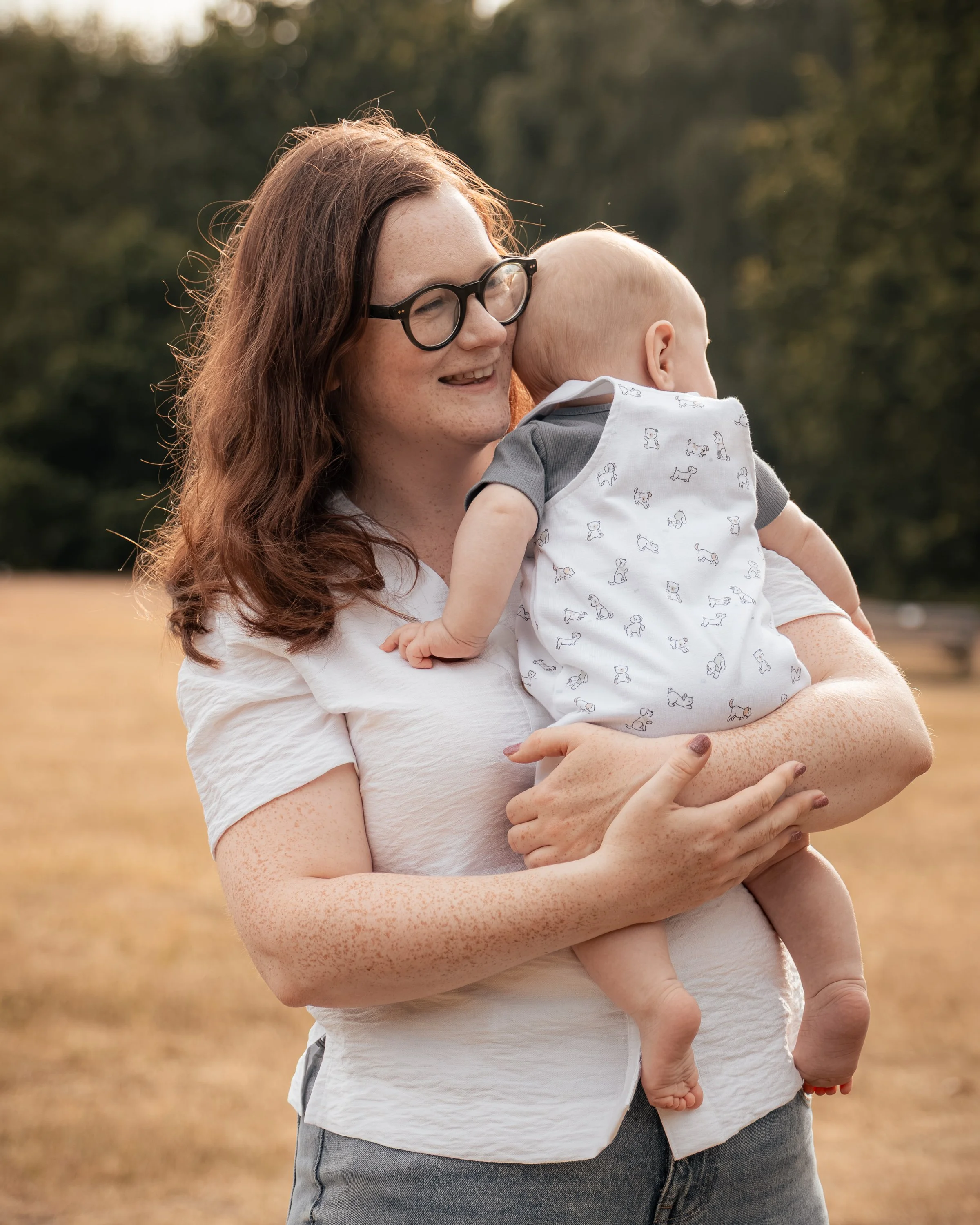 A woman with red hair and glasses holding a baby in an outdoor field with trees in the background.