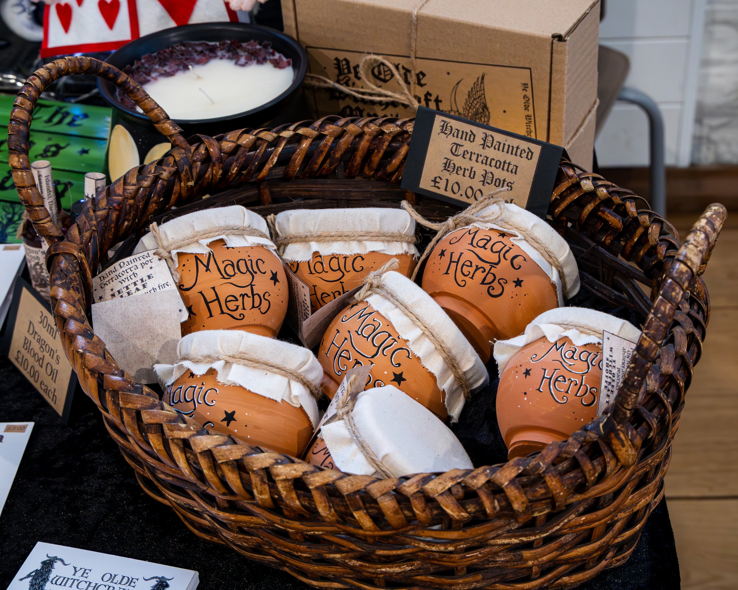 A wicker basket filled with jars of hand painted terracotta herb pots. A sign indicates they are priced at 10 pounds each. In the background, there is a black bowl with white wax and dried herbs and a cardboard box. Additional signs and items are vis