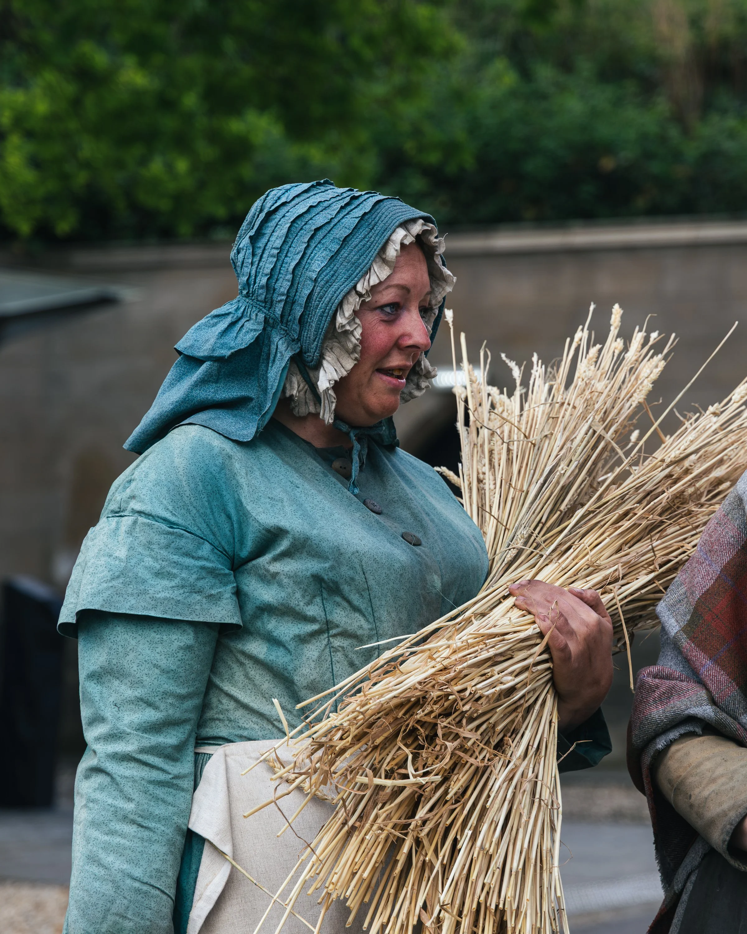 A woman with a towel on her head and a blue jacket holds a large bundle of straw or wheat stalks while talking to another person outside, with greenery in the background.