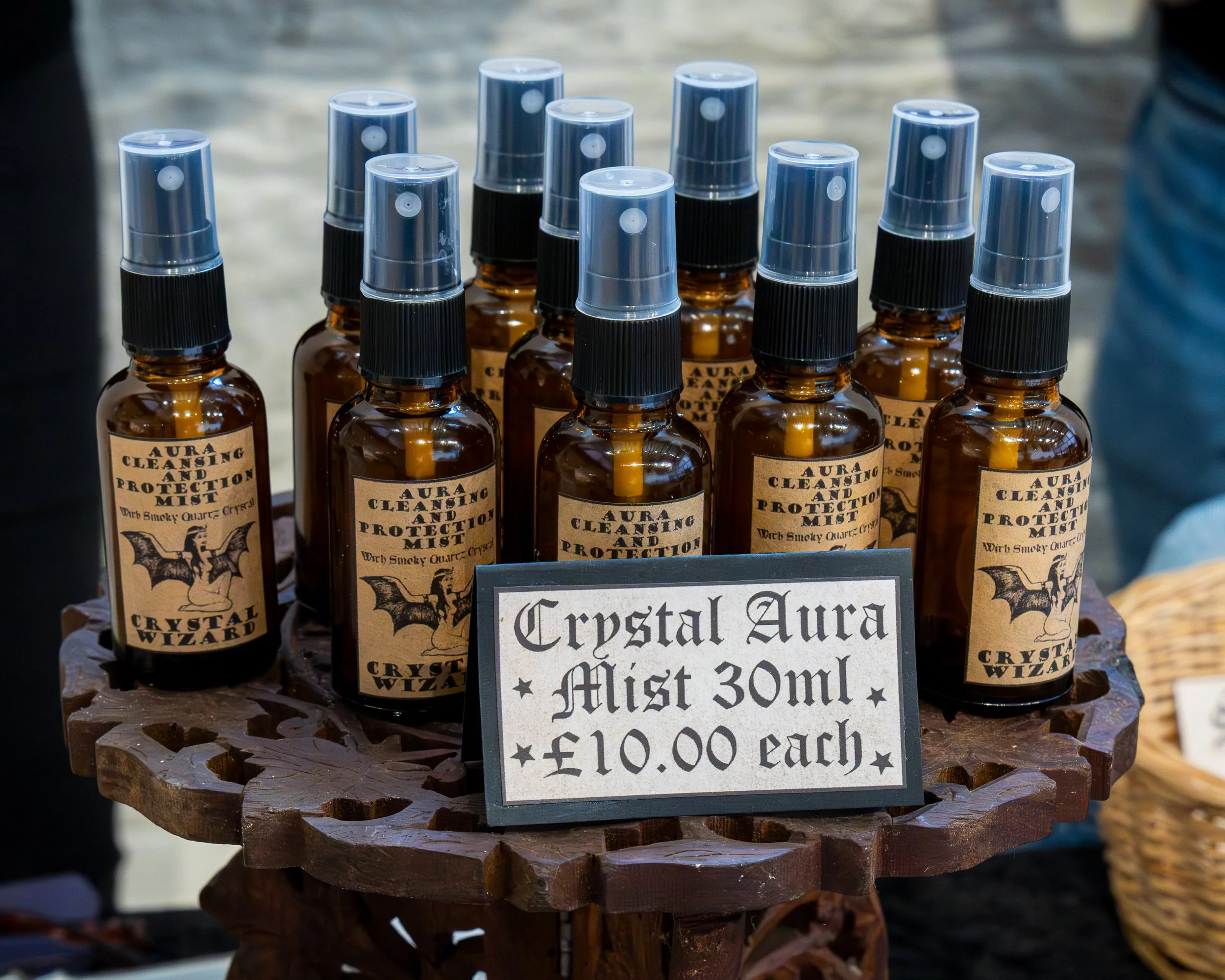 A display of small amber glass spray bottles with black spray nozzles and transparent caps. The bottles are labeled 'Aura Cleansing and Protection Mist,' priced at 10 dollars each. A sign in front details the product as 'Crystal Aura Mist 30ml' and m