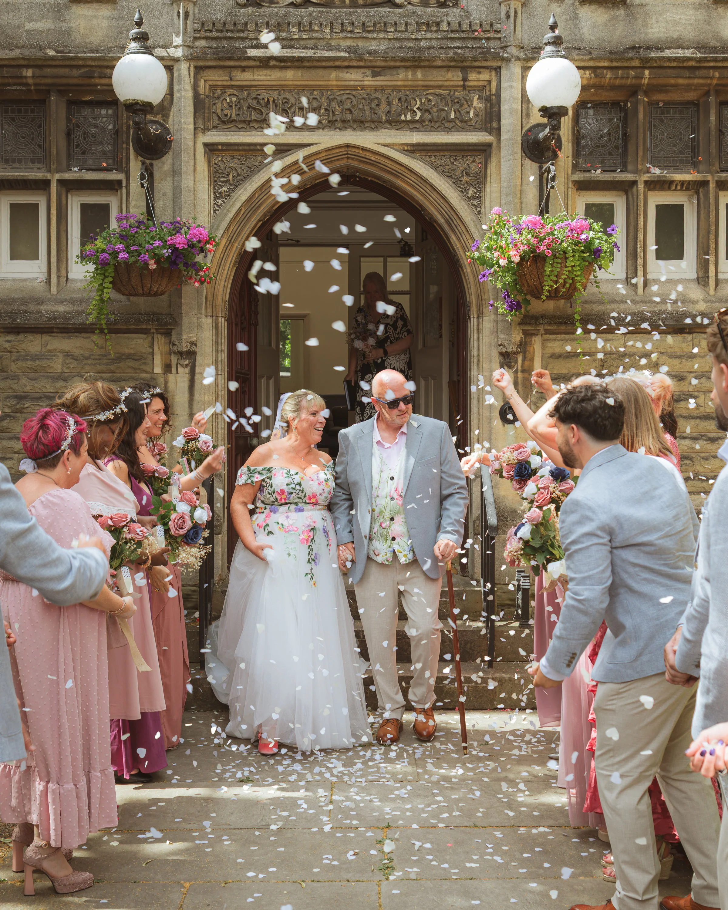 A festive outdoor wedding celebration with guests throwing flower petals or confetti in front of an ornate stone building entrance. The bride and groom are smiling, surrounded by friends dressed in pastel and floral attire