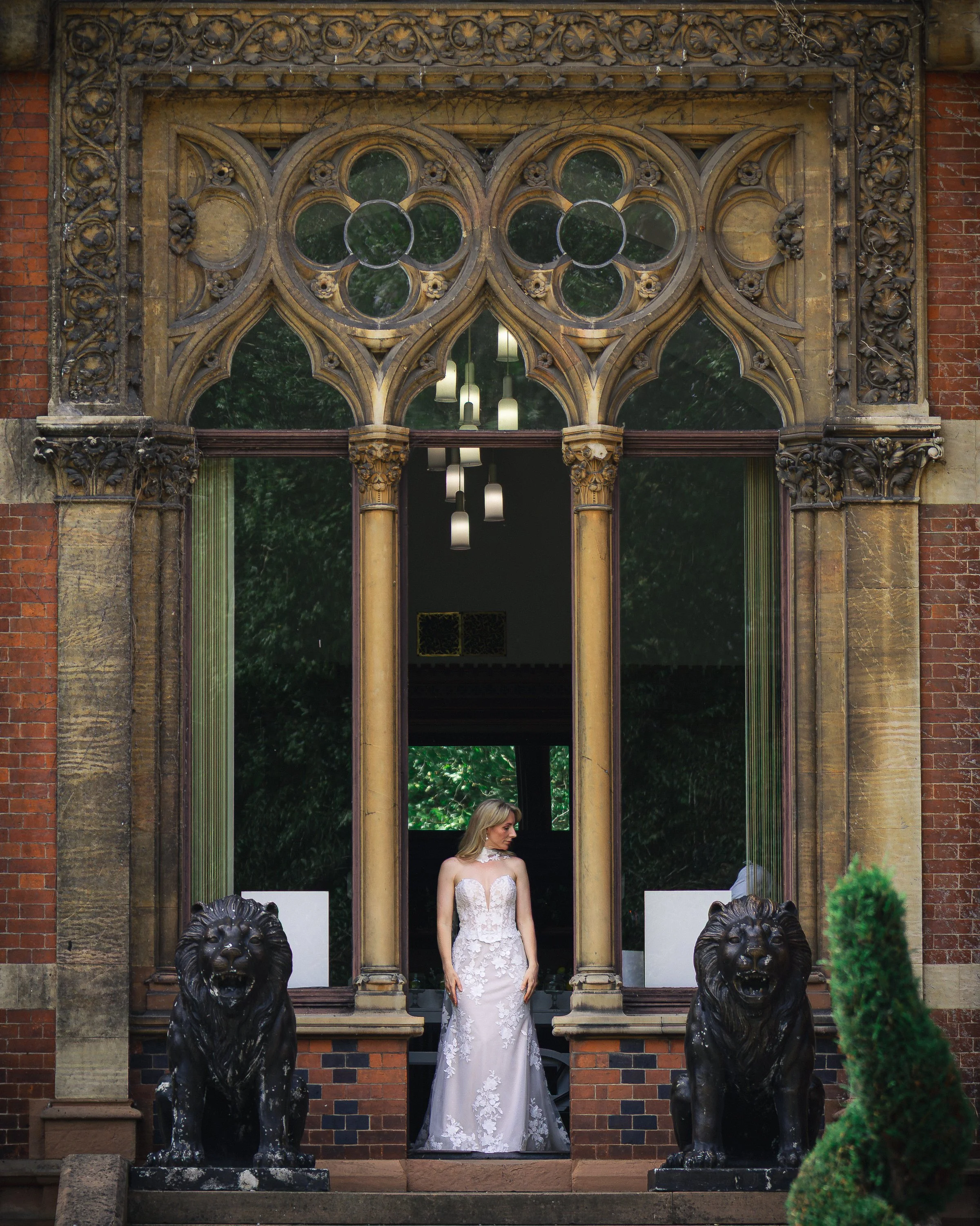 A woman in a white lace wedding dress standing in front of a large decorative window with stone carvings and two lion statues at the base, outside a brick building. The Renaissance at Kelham Hall