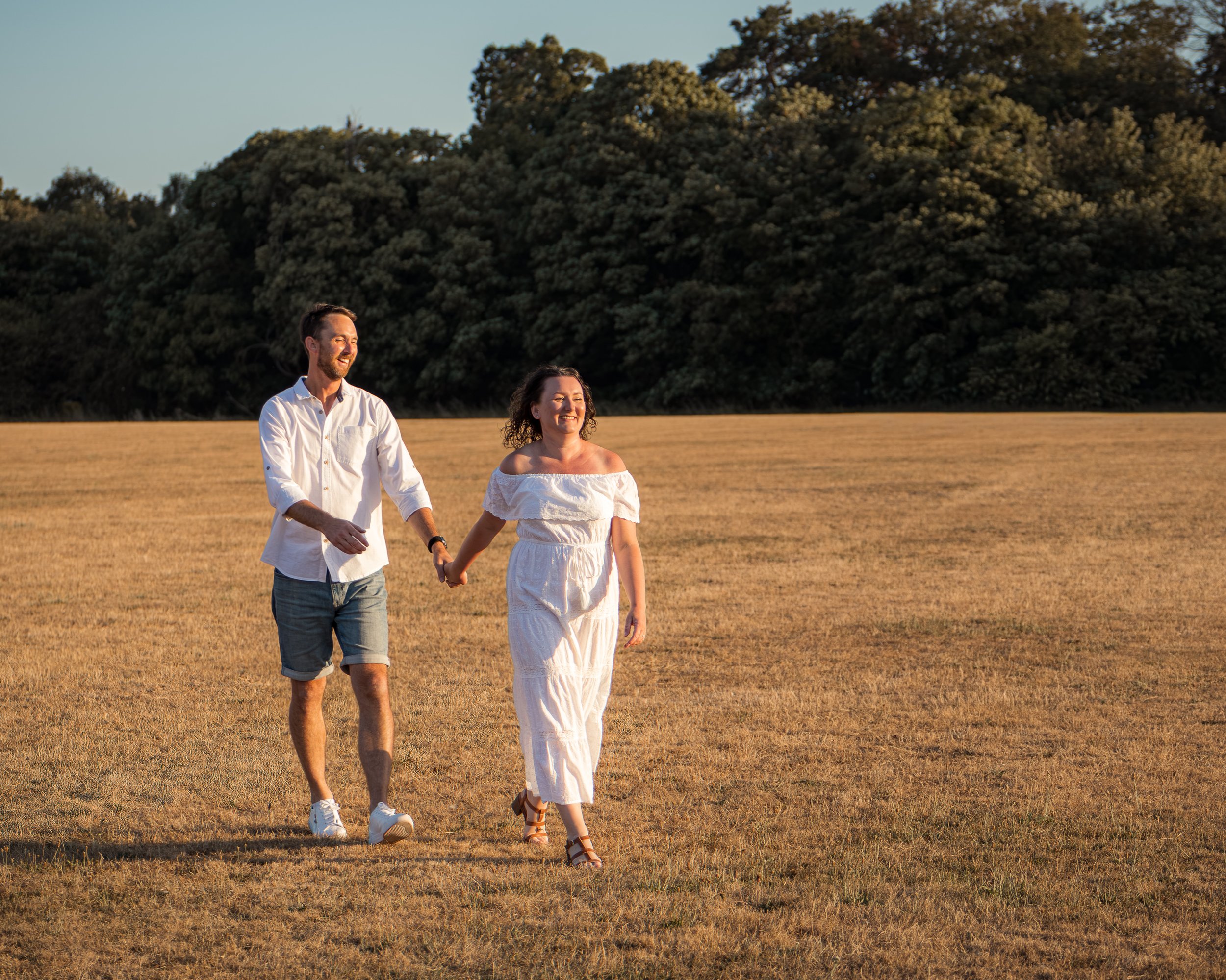 A couple is walking outdoors on a grassy field with a large forest of trees in the background, enjoying the sunlight and smiling.