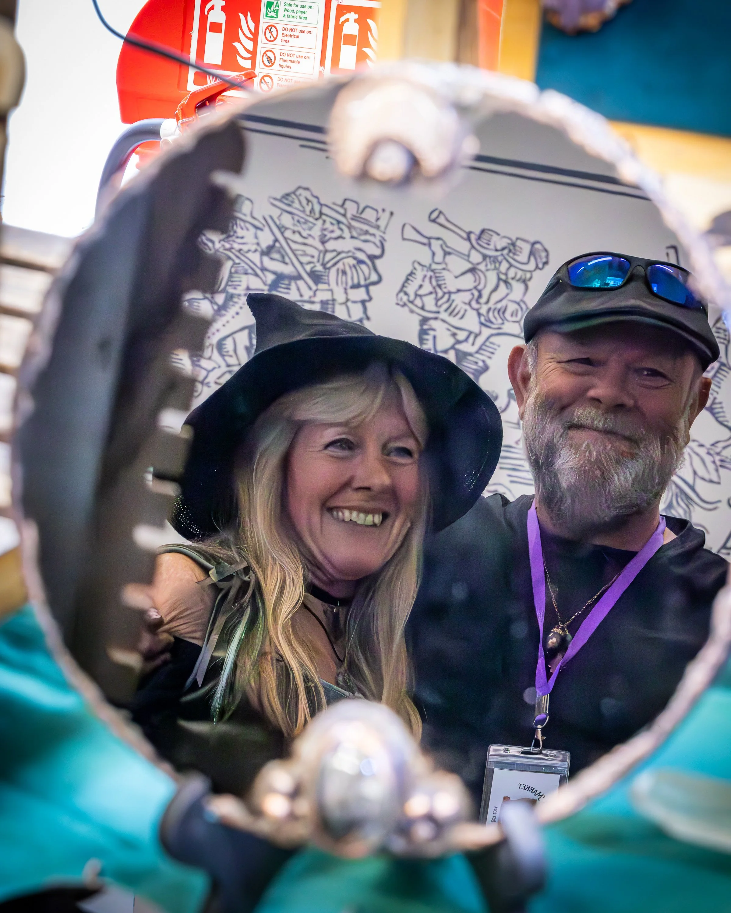 Smiling woman and man taking a selfie using a camera or phone through a hole in a metallic or reflective surface. The woman has long blonde hair and wears a wide-brimmed hat, while the man has gray hair, a beard, and glasses with blue tinted lenses. 