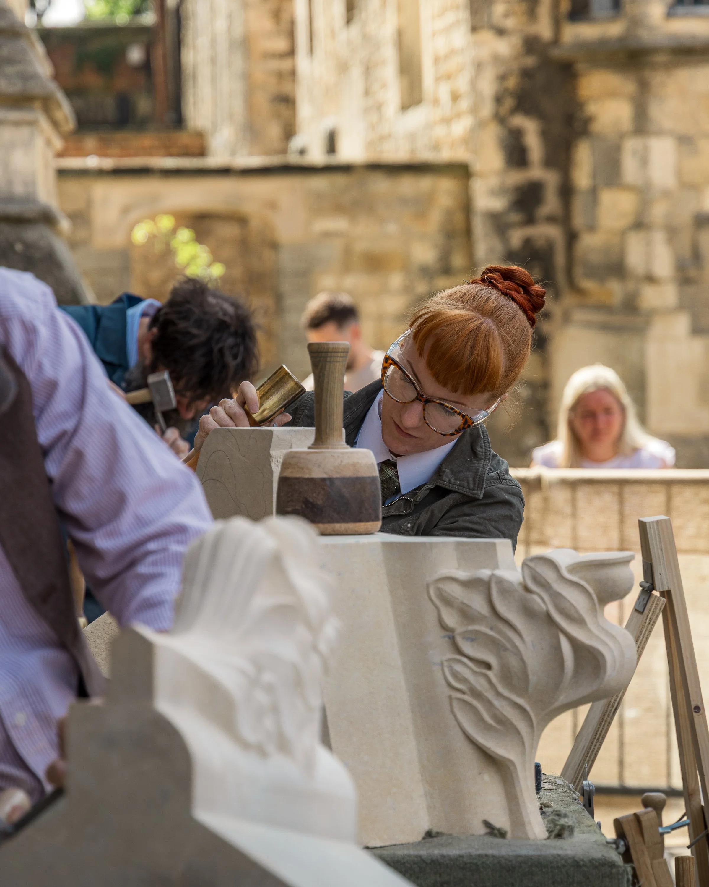 People working on a stone sculpture outdoors, with a historical stone building in the background.