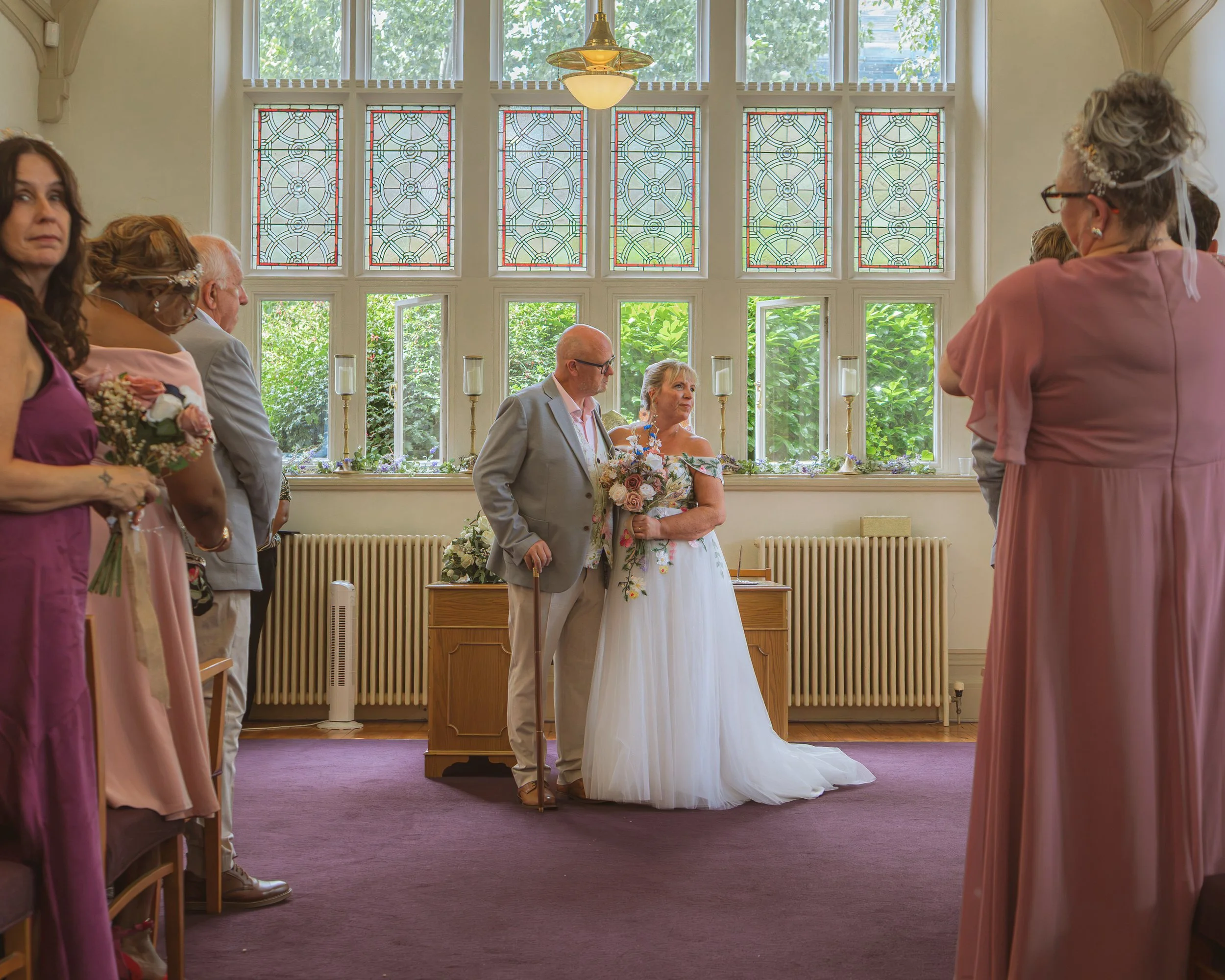 A wedding ceremony taking place inside a room with large windows and stained glass panels. The bride and groom stand at the altar, surrounded by guests. The bride is holding a bouquet, and the groom is wearing a light gray suit. The room has purple c