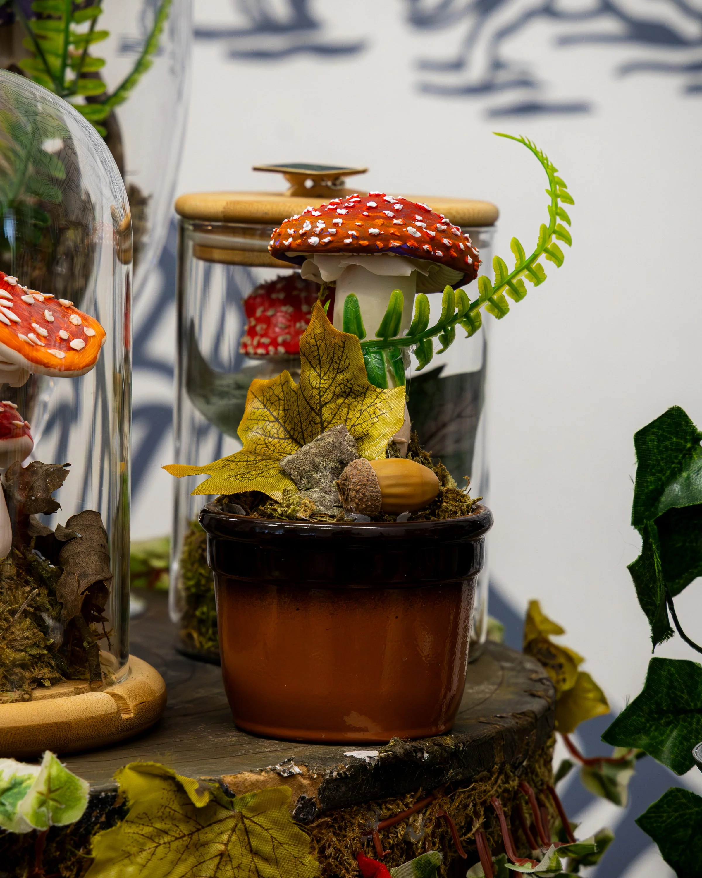Decorative mushroom-shaped ornament with a red, white-spotted cap and a white stem situated in a brown pot, surrounded by artificial leaves and other natural-looking elements.
